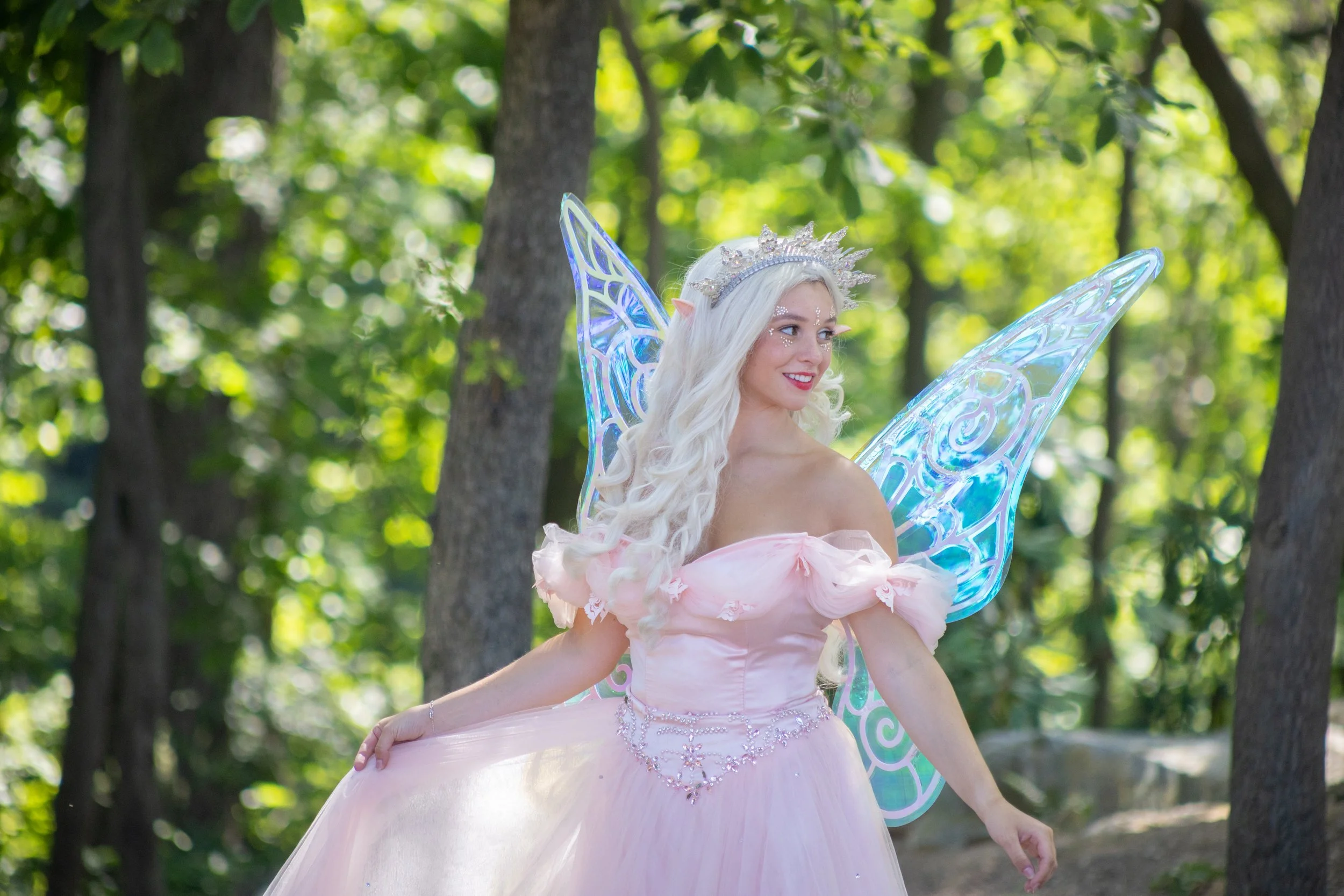 A woman dressed as a fairy with long white hair, blue fairy wings, and a pink gown, standing outdoors in a wooded area.