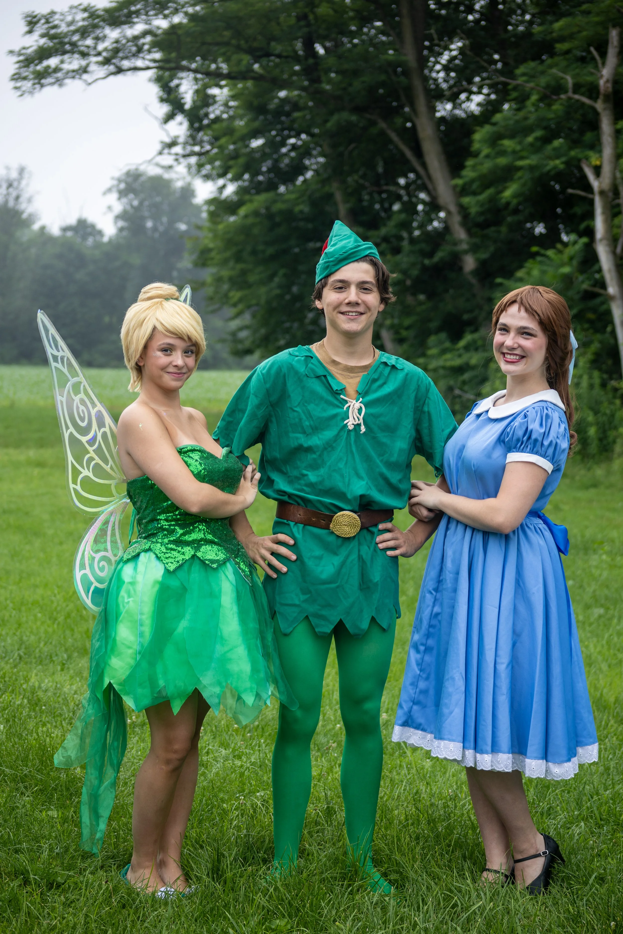 Three people dressed as a Tinkerbell, Peter Pan and Wendy standing in a field with trees in the background. 