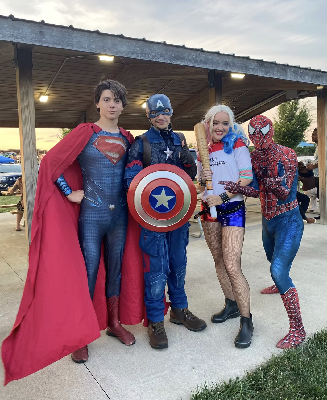 Group of five people dressed as superheroes and comic characters, inspired by Superman, Captain America, Harley Quinn, and Spider-Man, standing outdoors near a pavilion.
