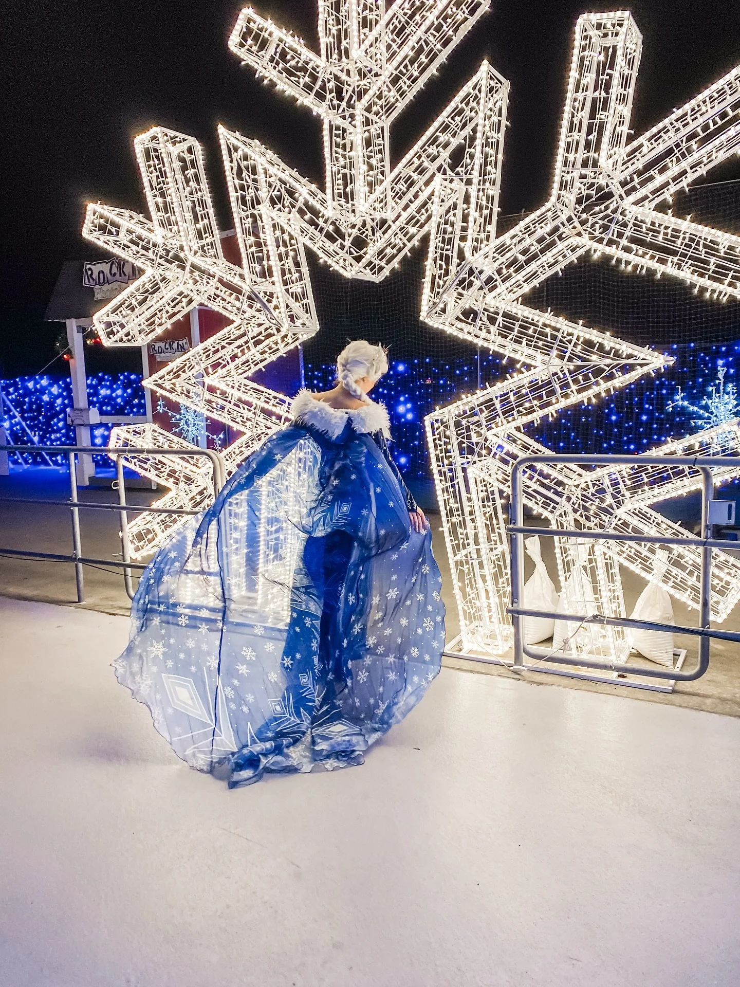 A person dressed as Elsa from Frozen in a blue gown with snowflake patterns, standing in front of lit-up Christmas star decorations and blue lights at night.