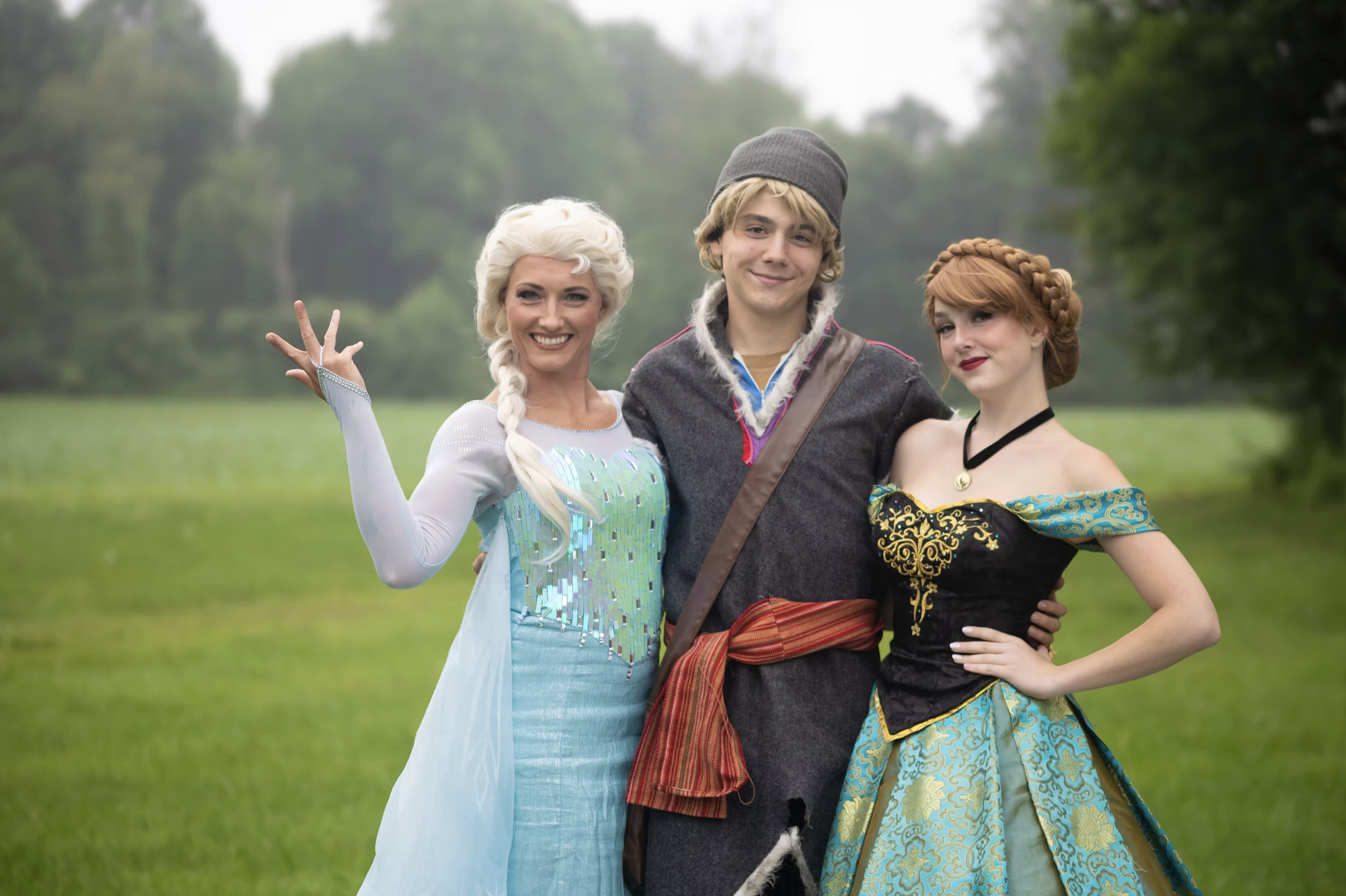 Three people dressed as characters from Frozen and a prince, standing outdoors on grass with trees in the background.