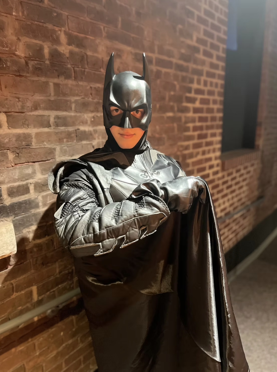 A person dressed as Batman, wearing a black Batman mask, a dark leather-like costume, and gloves, standing against a brick wall.