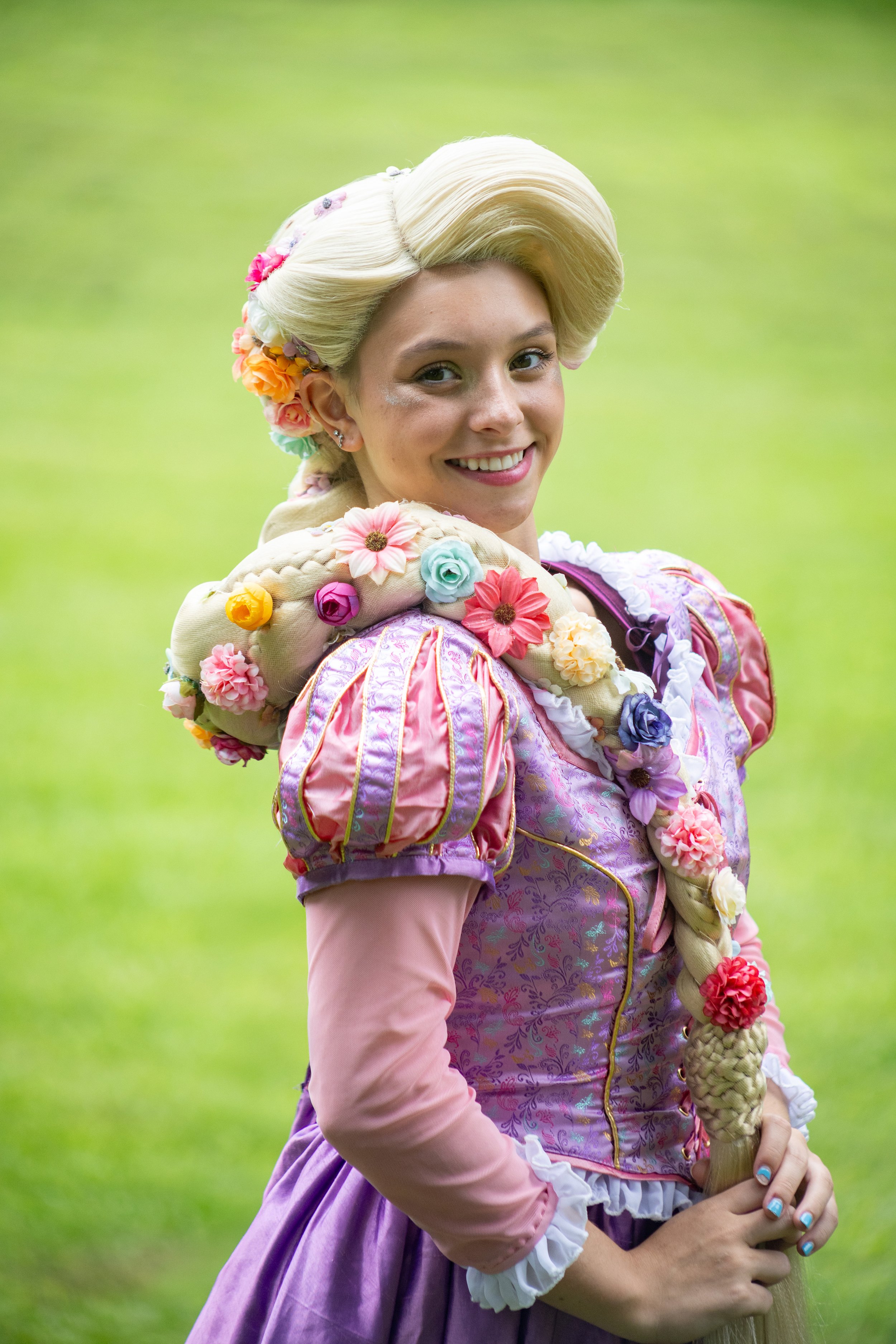 Woman dressed as Rapunzel with a long, braided hair adorned with colorful flowers, standing outdoors on a grassy background.
