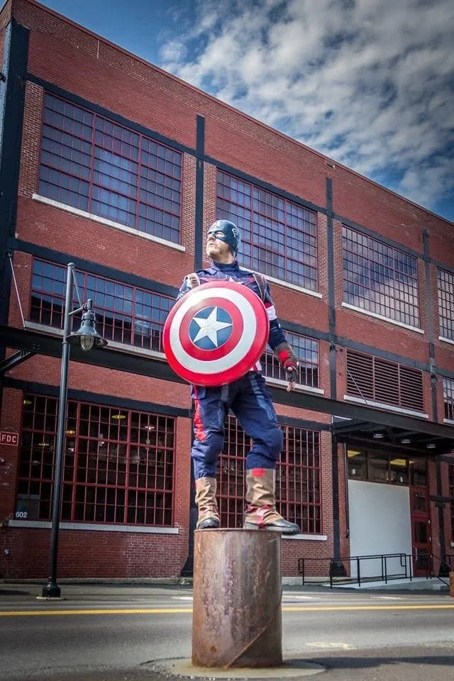 Inspired by Captain America standing on a metal pillar in front of a brick building, holding a shield with a star, in an urban setting with a cloudy sky.