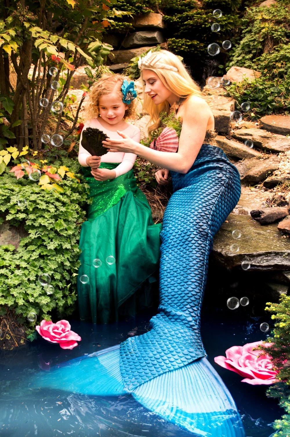 A woman and a young girl dressed as mermaids sitting by a small pond in a lush garden, looking at a seashell together surrounded by green plants, rocks, and floating pink flowers.