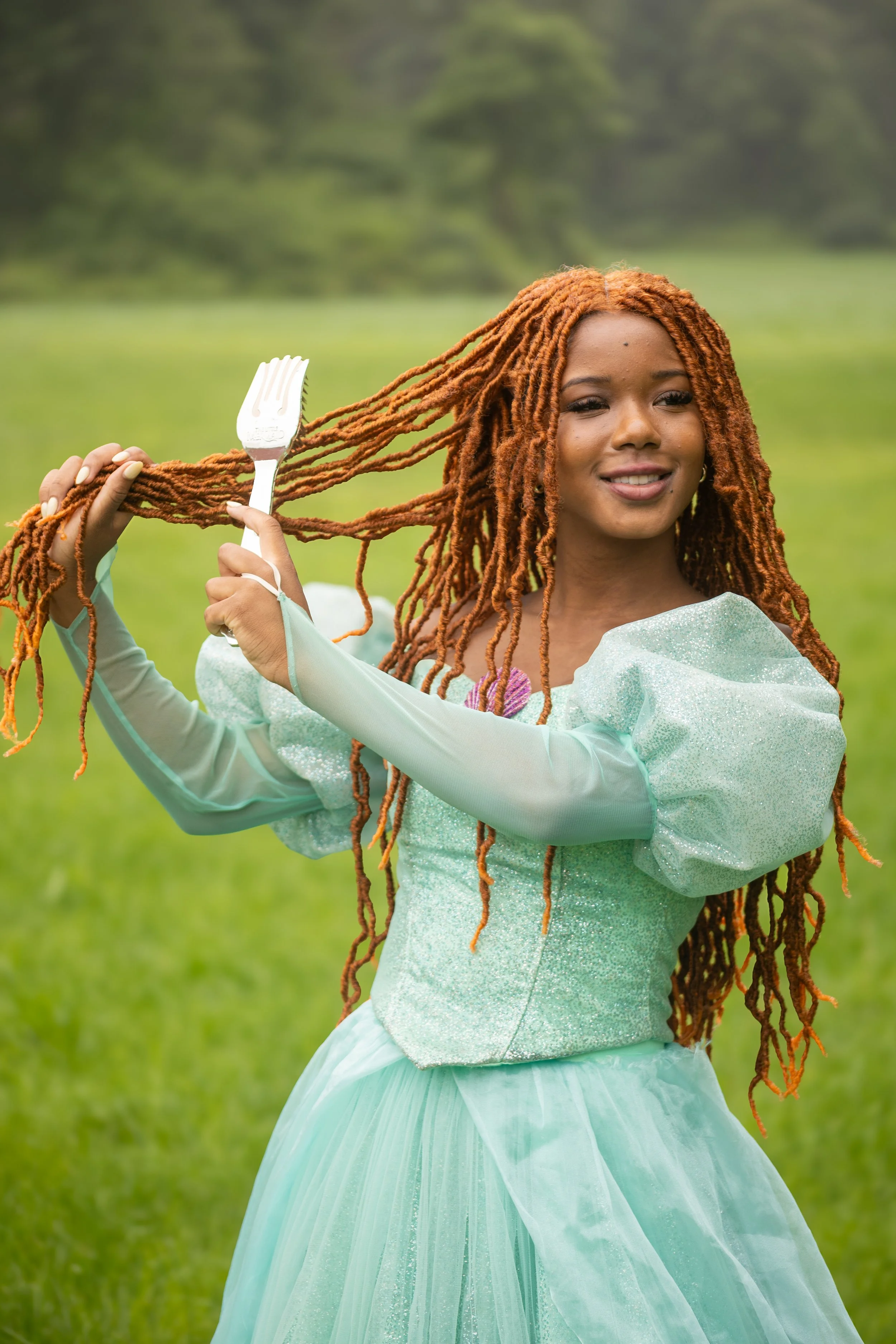 A young woman with long, orange, twisted dreadlocks outdoors in a grassy area, holding a silver fork in her hand, wearing a light green, sparkly, puffy-sleeved gown and smiling.