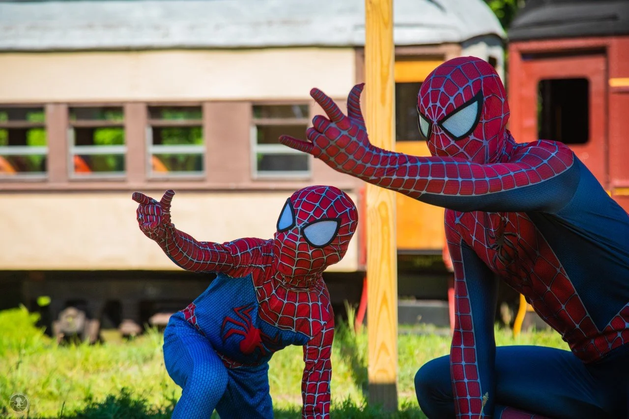 Two children dressed as Spider-Man posing outdoors near a wooden structure, with a train in the background.
