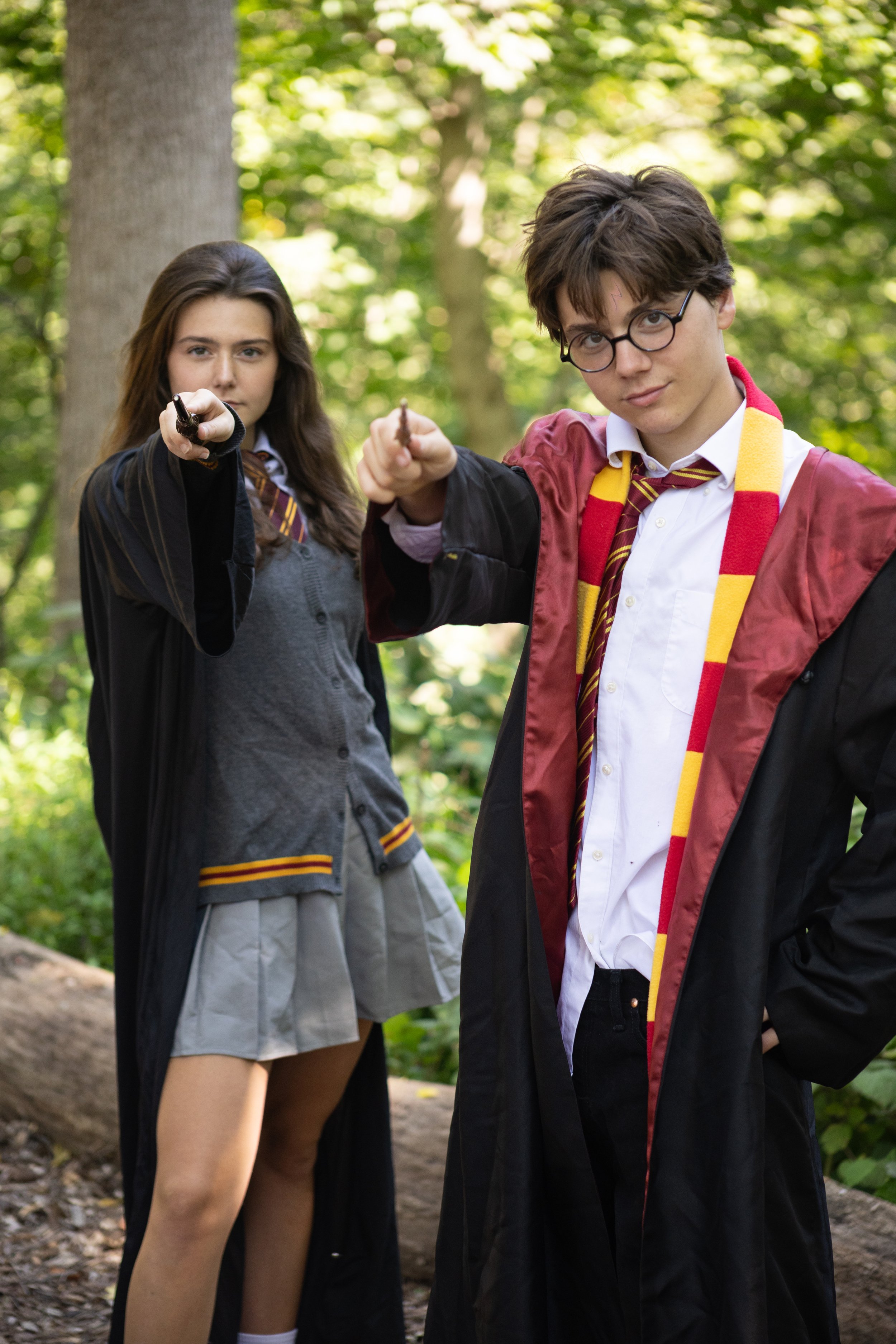 Two teenagers in Harry Potter costumes, a girl and a boy, standing outdoors in a wooded area, both pointing wands towards the camera.