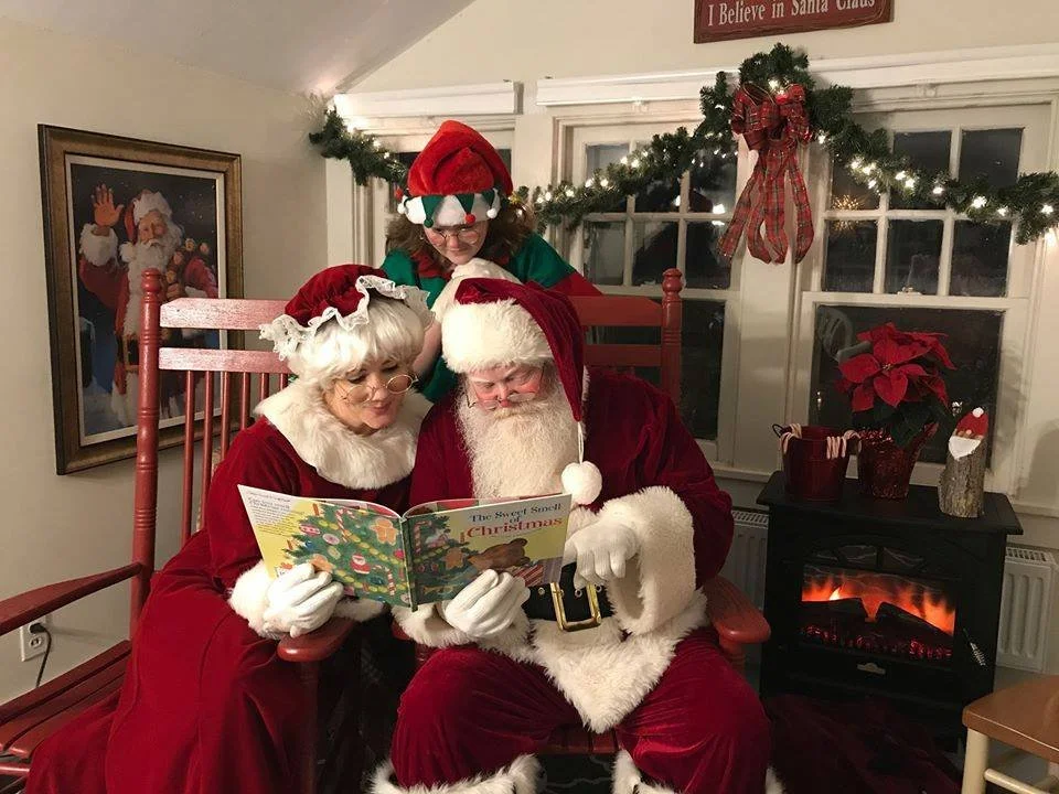 Three people dressed as Santa Claus, Mrs. Claus, and an elf are gathered around reading a Christmas book titled 'The Sweet Smell of Christmas,' in a festive living room with Christmas decorations and a fireplace.