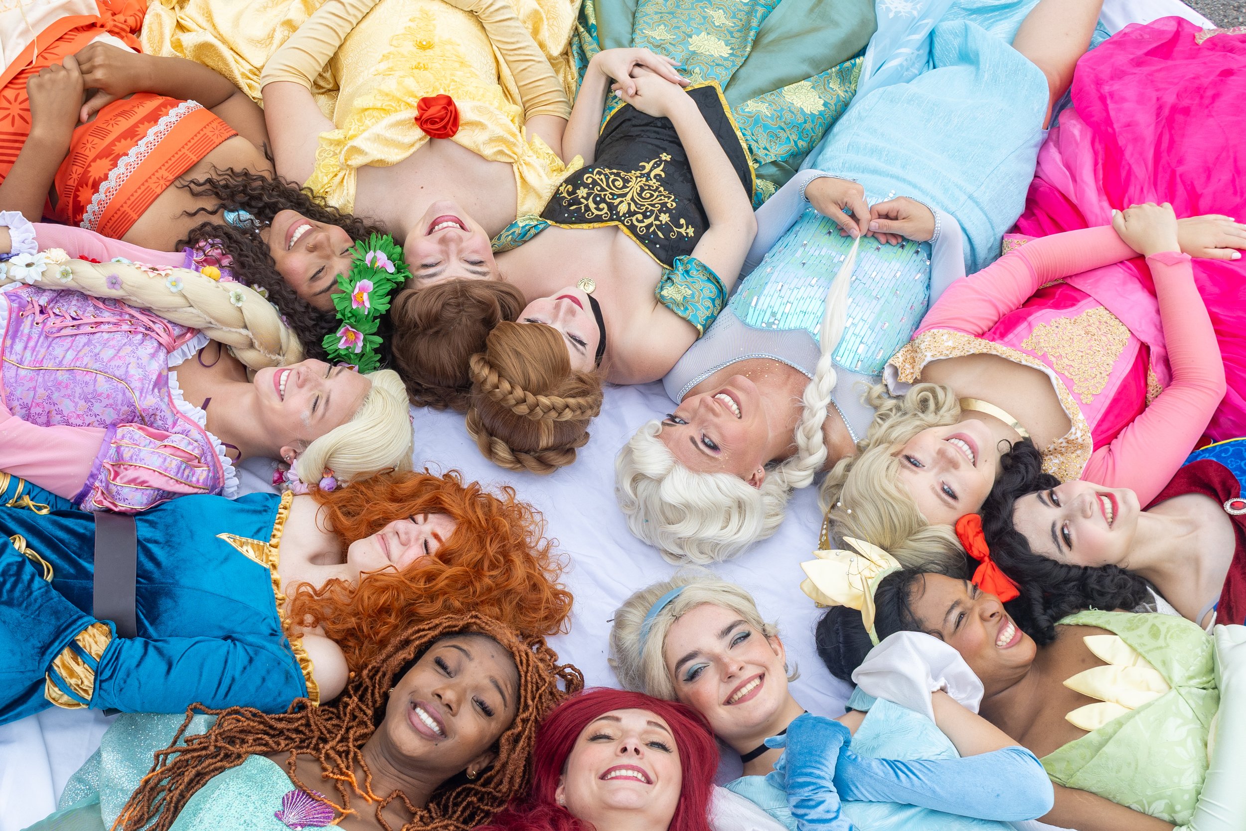 Group of women dressed as Disney princesses lying in a circle on a white surface, smiling and relaxing.