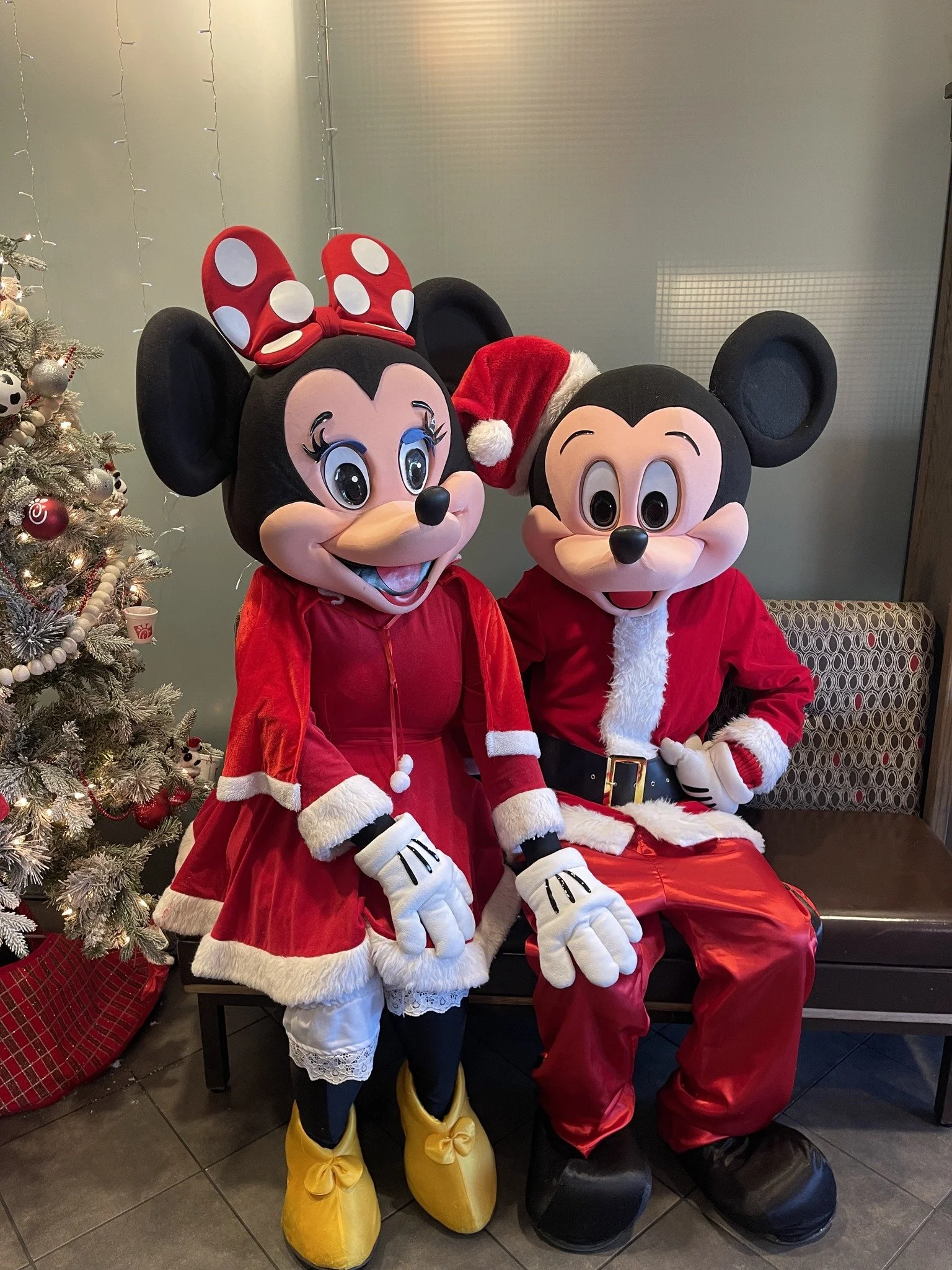 Minnie Mouse and Mickey Mouse dressed in Christmas costumes, Minnie in a red dress with white fur trim, yellow shoes, and a red bow with white polka dots, Mickey in a Santa suit, sitting on a bench with a Christmas tree decorated with ornaments and lights beside them.