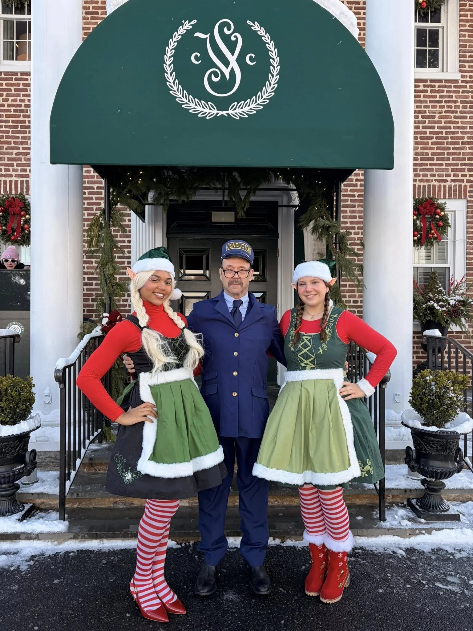 Two women dressed as Christmas elves and a man dressed as a train conductor stand in front of a decorated building entrance with Christmas decorations and snow on the ground.