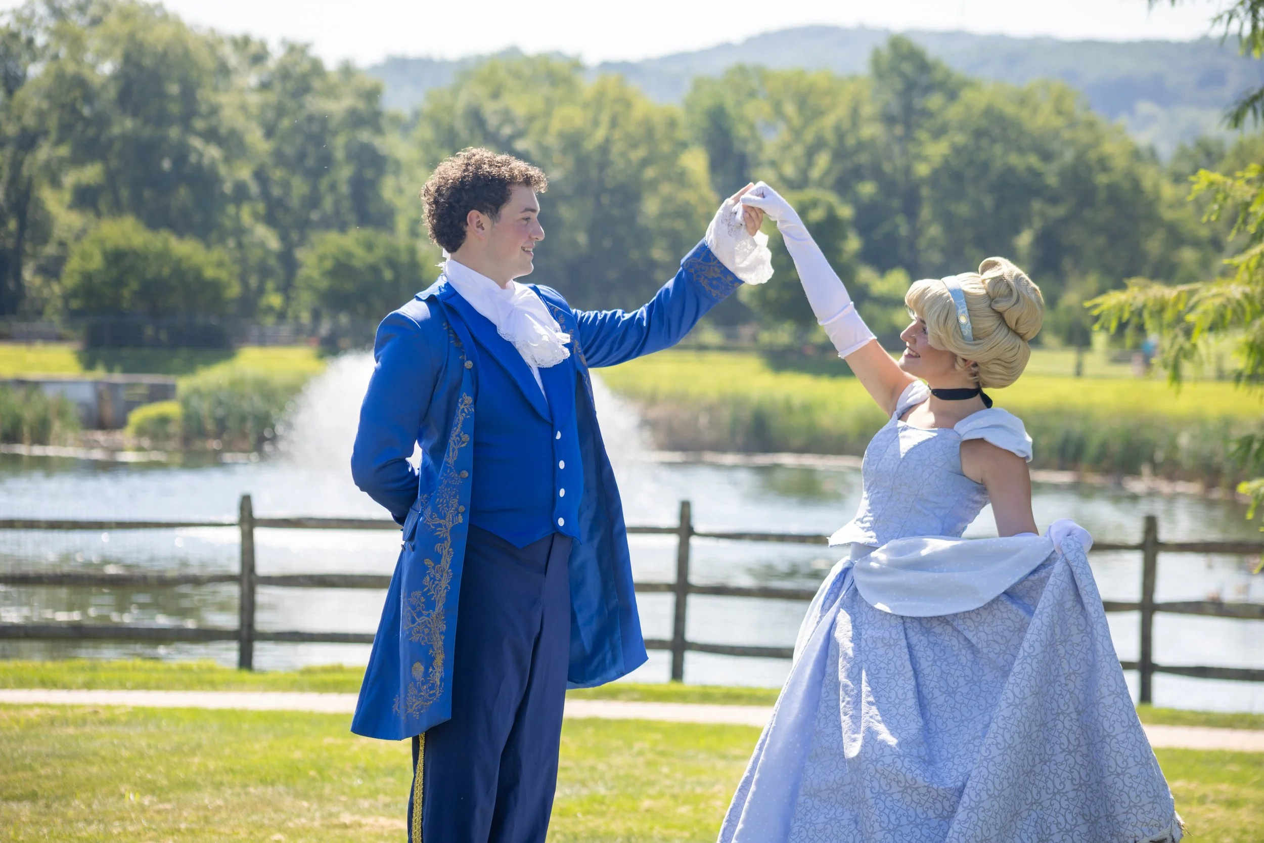 Couple dressed in historical costumes dancing outdoors by a pond with a wooden fence, green trees, and hills in the background.