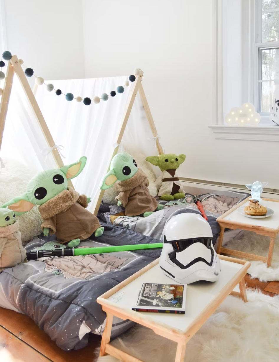 Children's bedroom decorated with Star Wars and Star Wars-themed plush toys, including Yoda and Grogu, along with Star Wars helmet, lightsaber toys, books, and themed decor.
