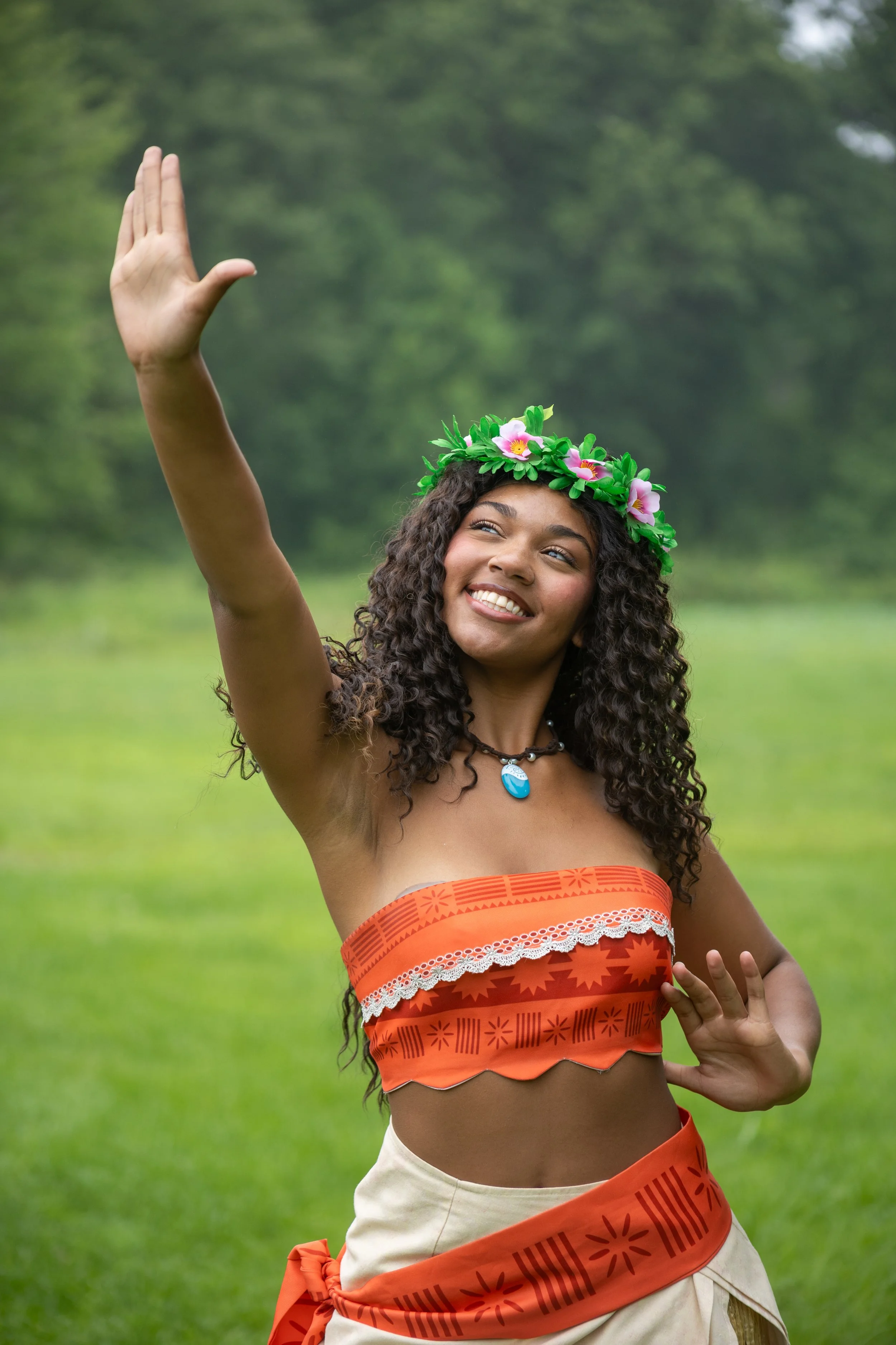 A woman with curly hair and a floral headband smiling and raising her right hand in a grassy outdoor setting.