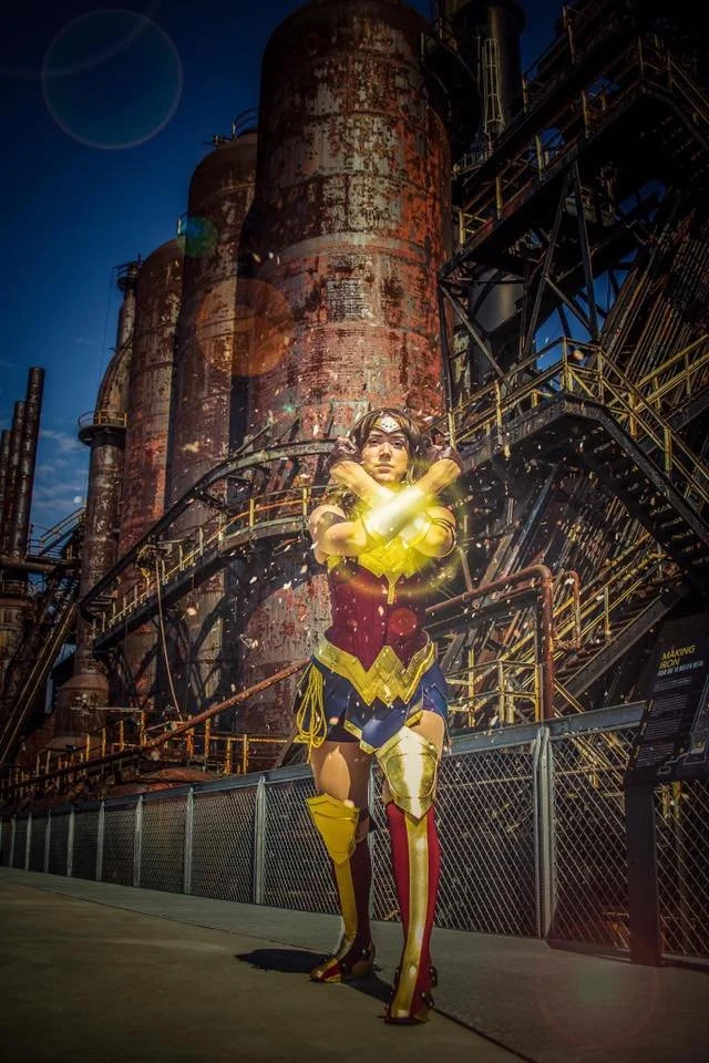 Inspired by Wonder Woman with glowing bracelets in front of an industrial factory with large rusted pipes and metal structures at sunset.