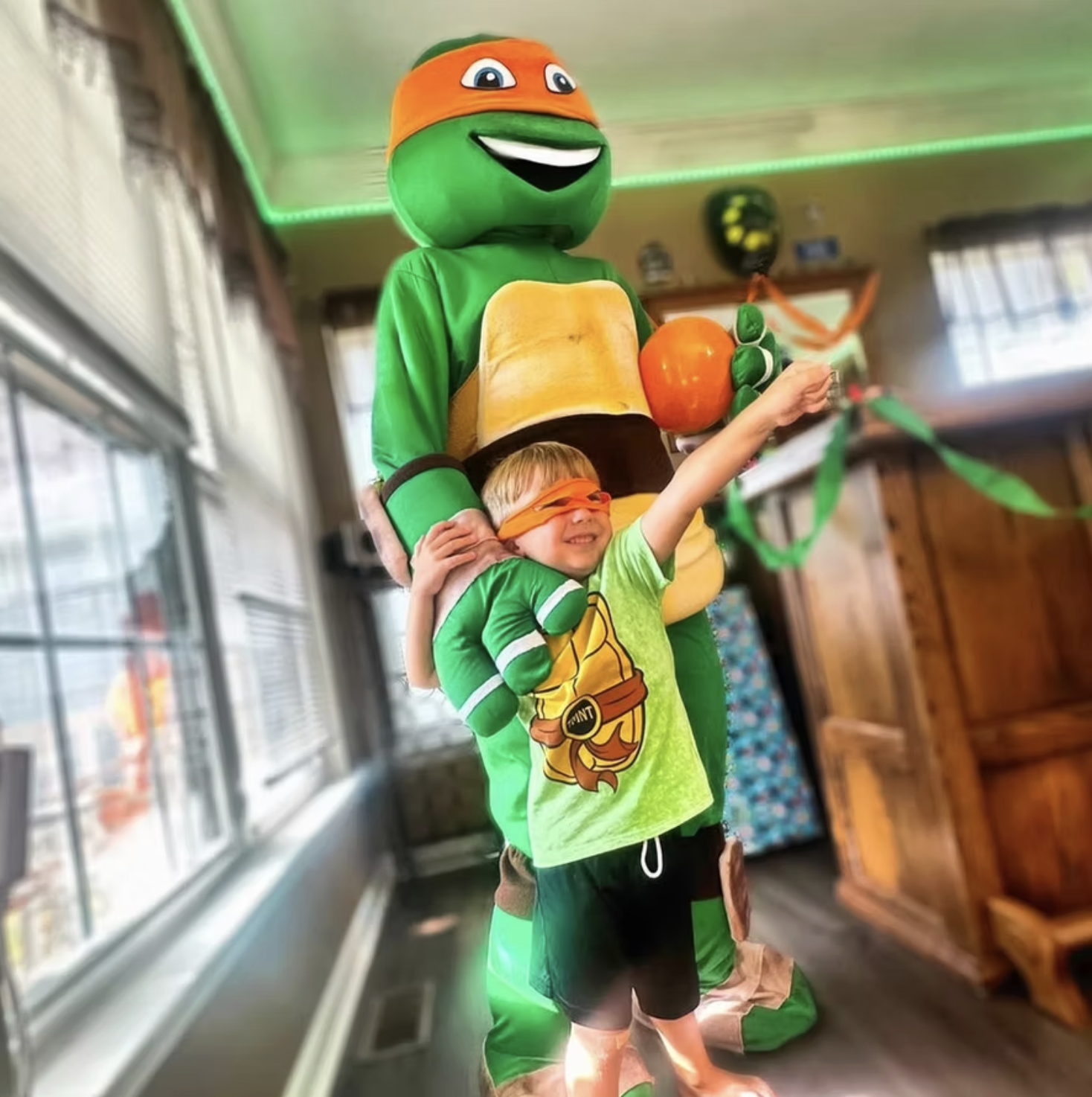 A child wearing an orange mask and green shirt is celebrating with a Teenage Mutant Ninja Turtles mascot dressed as a turtle, holding an orange ball, during a birthday party.