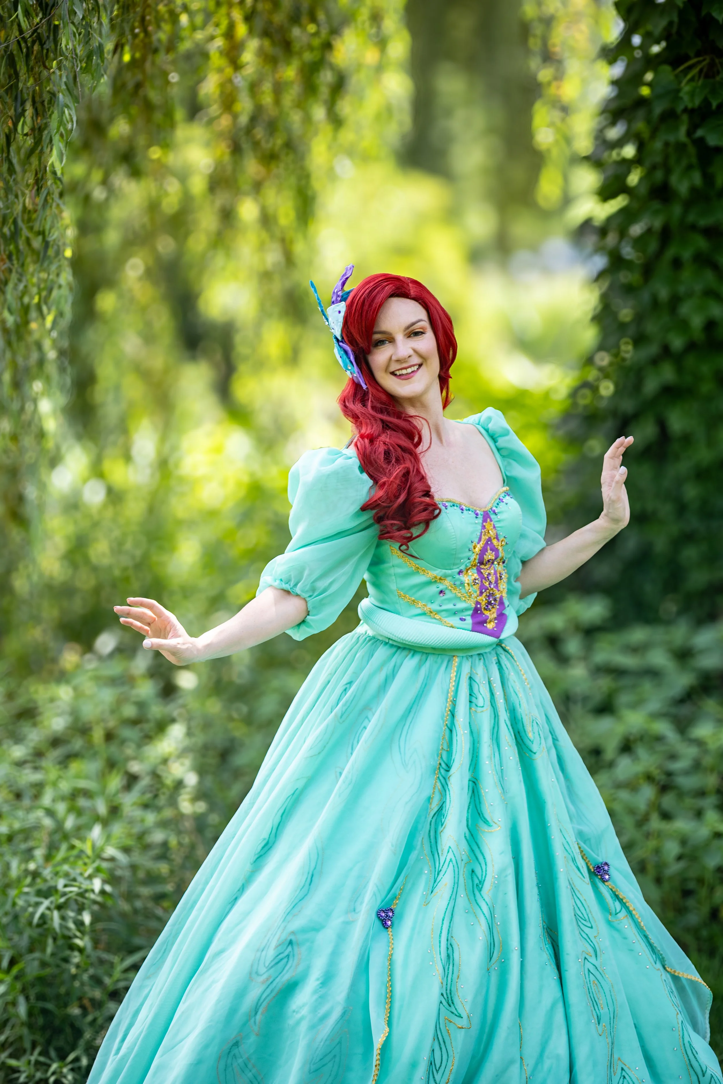 A woman with long red hair wearing a teal princess dress with puffed sleeves and intricate detailing, standing outdoors among green foliage, smiling at the camera with her arms slightly raised, inspired by Princess Ariel.
