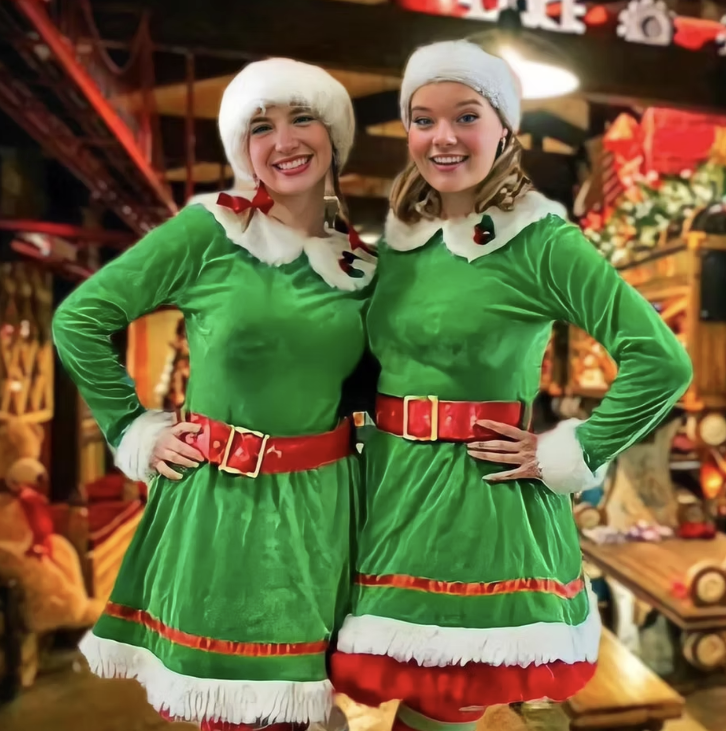 Two women dressed as Christmas elves in green velvet dresses with white and red accents, white hats, and smiling for a photo in a festive indoor setting decorated for Christmas.