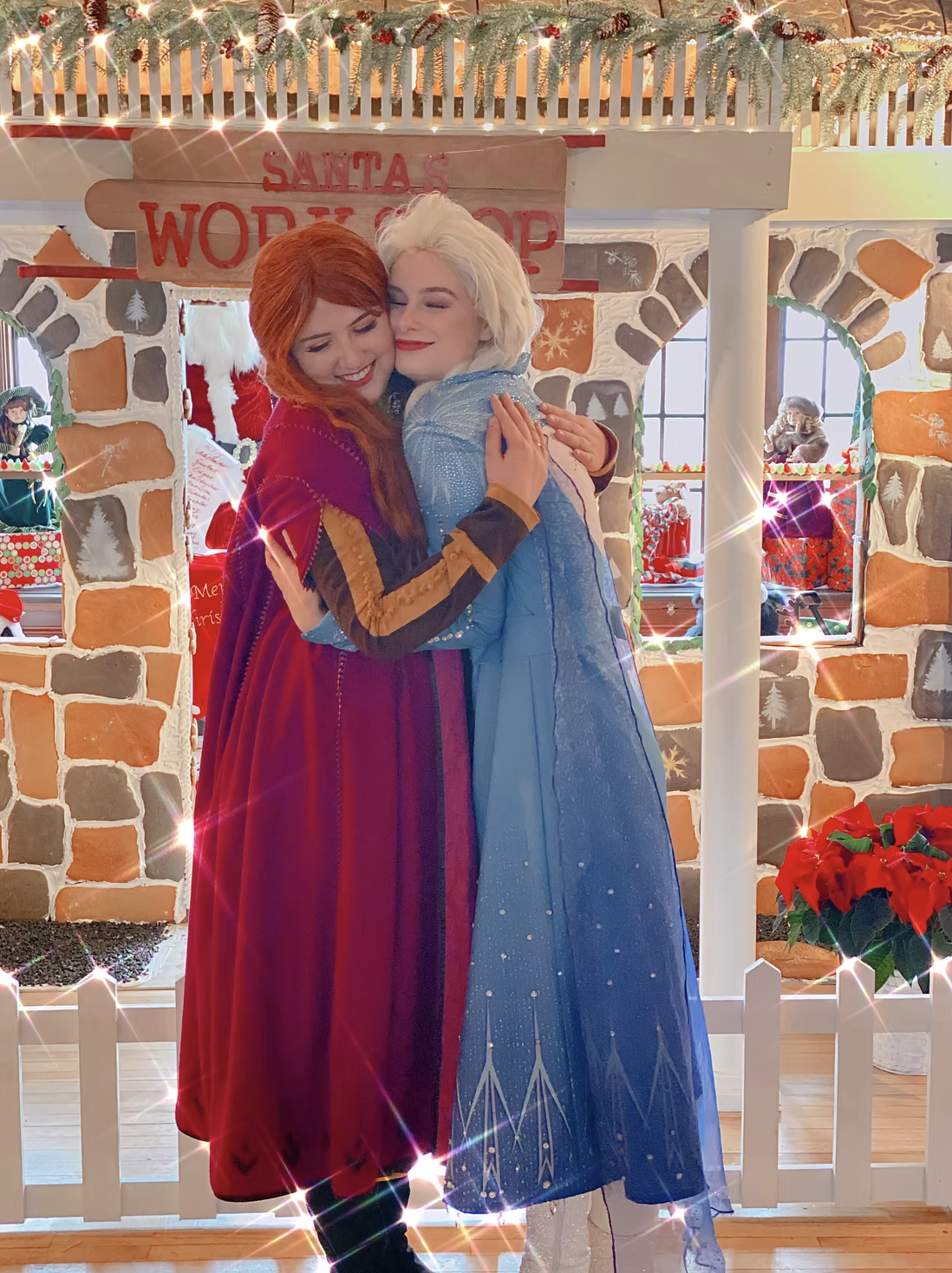Two women dressed as Disney characters, Anna and Elsa from Frozen, hugging in front of a Christmas-themed backdrop with a sign that reads 'Santa's Word Shop.' They are smiling with closed eyes amidst festive decorations including a poinsettia, Christ