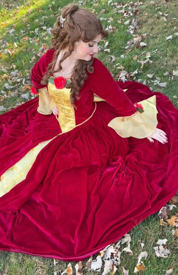 A woman dressed as Belle from Beauty and the Beast, in a red and yellow princess gown sitting on the grass among fallen leaves.