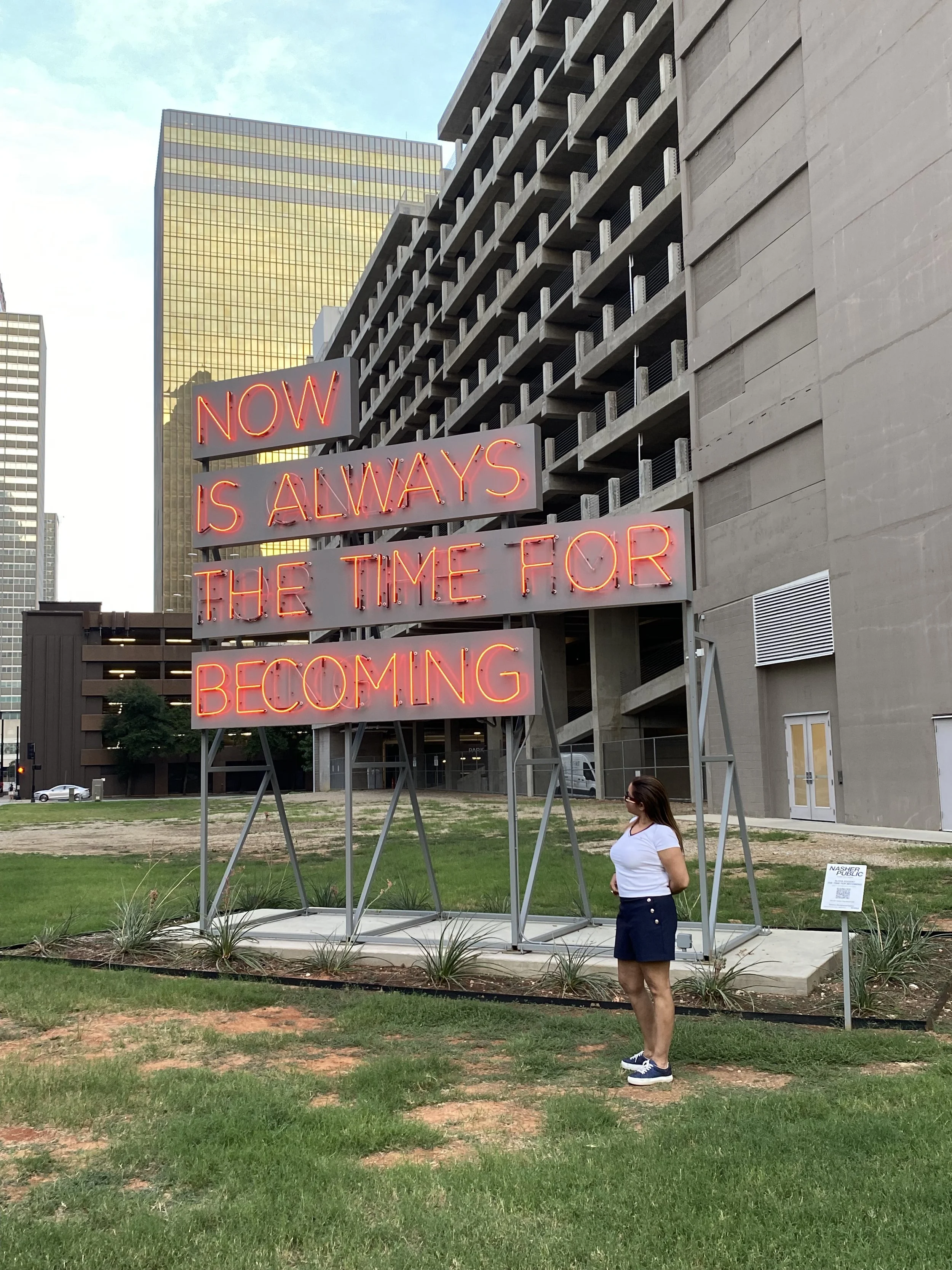 Neon sign in an urban area reads, "Now is Always the Time for Becoming." A woman in a white t-shirt, navy shorts, and sneakers stands nearby, looking at the sign, with modern buildings in the background.