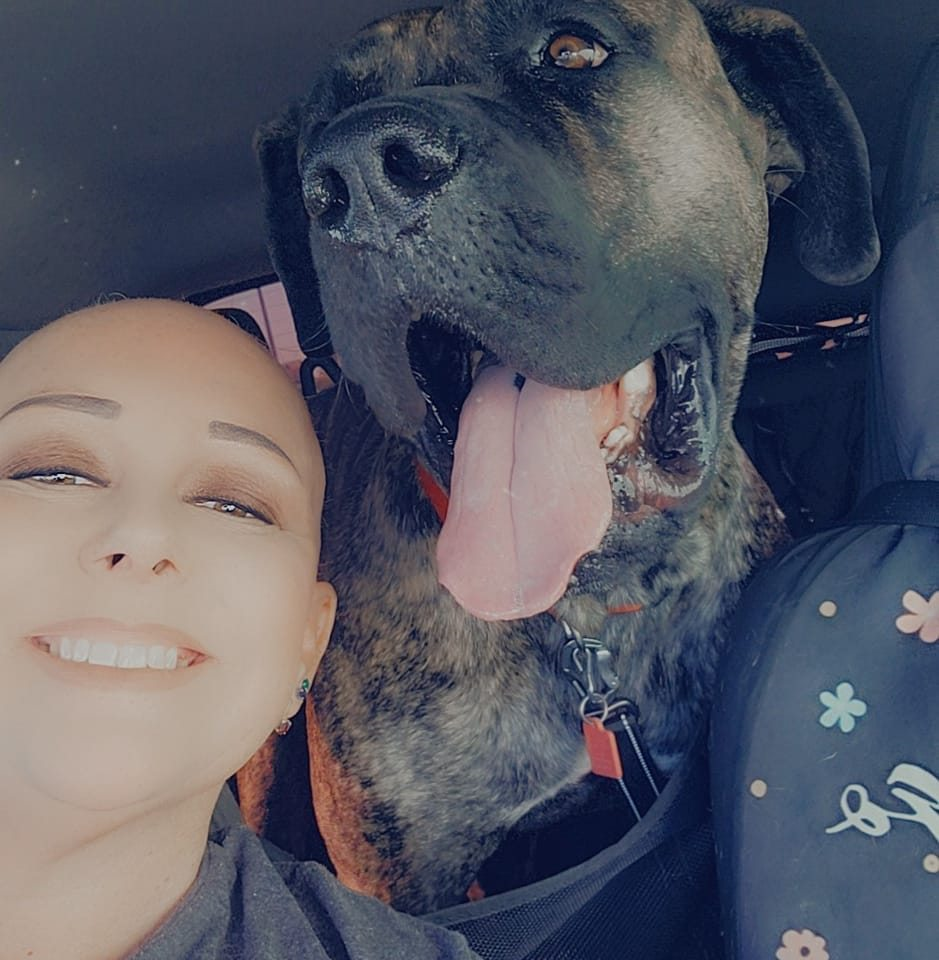 A woman smiling with a happy expression, next to a large, brindle-coated dog with its mouth open and tongue hanging out, sitting in a car.