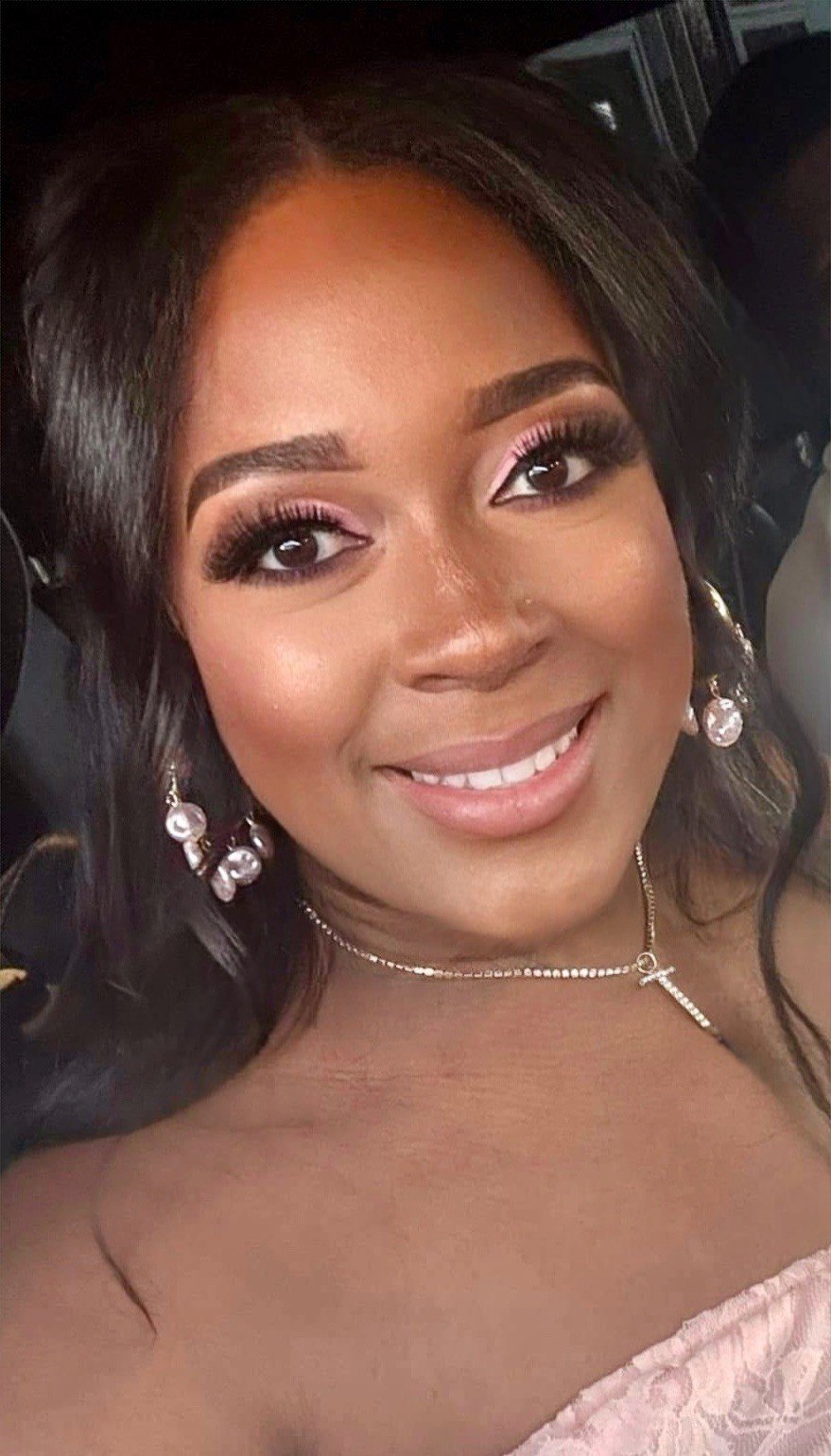Close-up of a smiling woman with dark hair, styled in loose waves, wearing makeup with pink eyeshadow and false eyelashes, pearl earrings, a delicate necklace with an cross, and an off-shoulder pink lace dress.