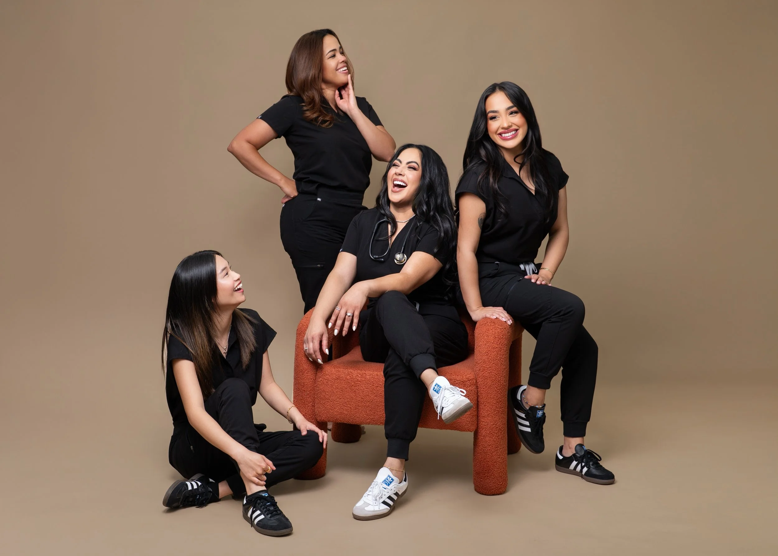 Five women, dressed in black clothing, are gathered together in a studio with a beige background. One woman is sitting on a rust-colored chair, smiling and wearing a stethoscope. Another woman is sitting on the floor, looking up and smiling at the se