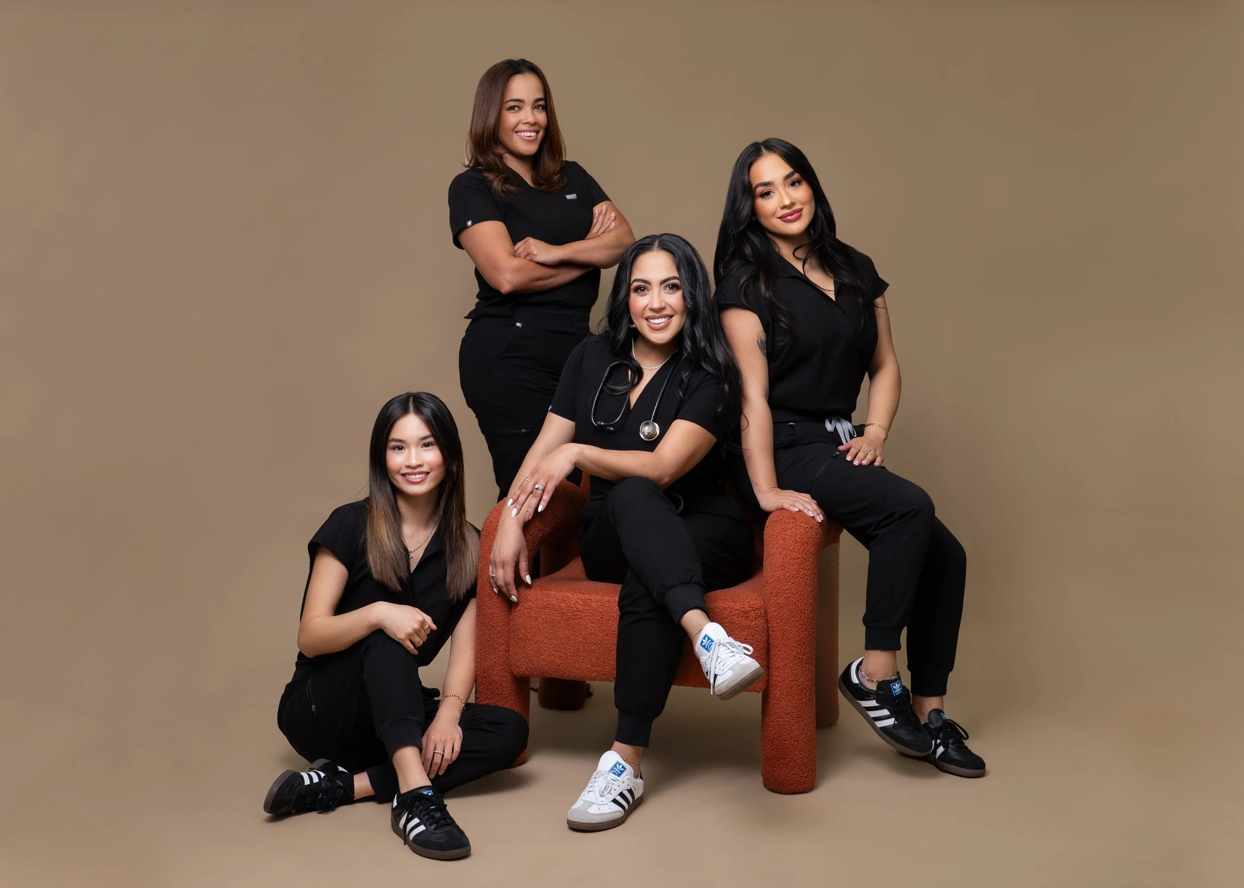 Group of five diverse women in black clothing posing together against a beige background, with one seated on an orange chair, one squatting, and three standing.