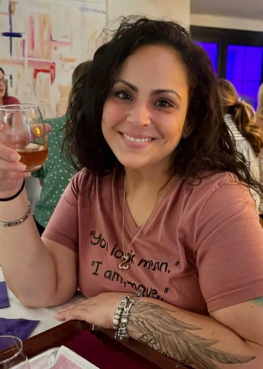 A woman with dark curly hair smiling at the camera while holding a glass of pink beverage at a social gathering.