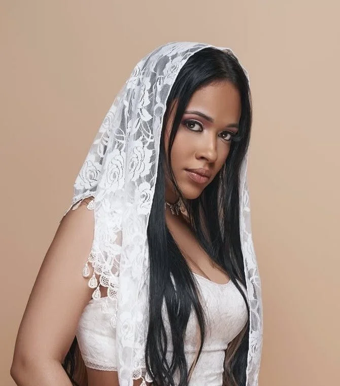 A woman with long black hair wearing a white lace head covering and a white top, posing against a beige background.