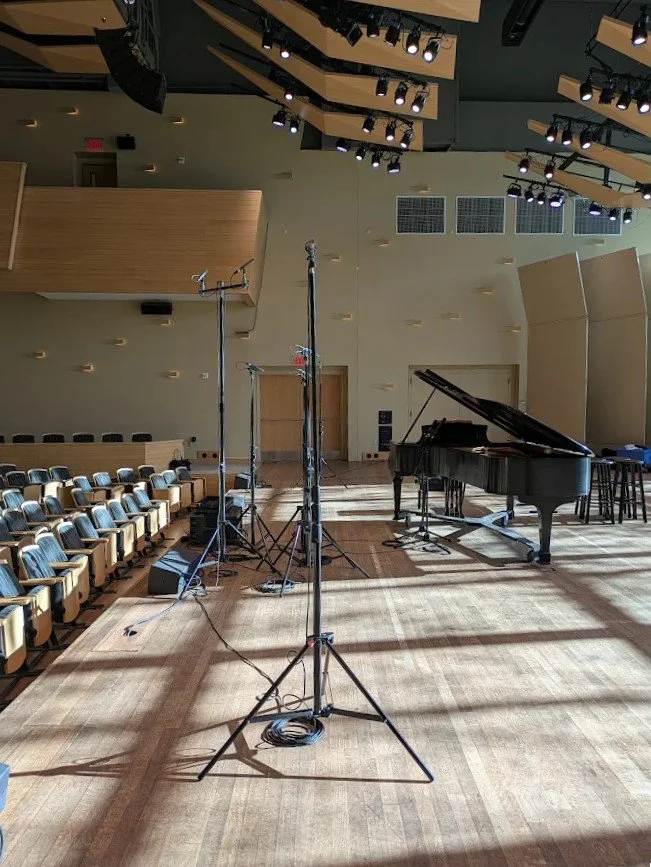Empty concert hall or auditorium stage with a grand piano, microphones, and audio equipment, ready for a performance or rehearsal.
