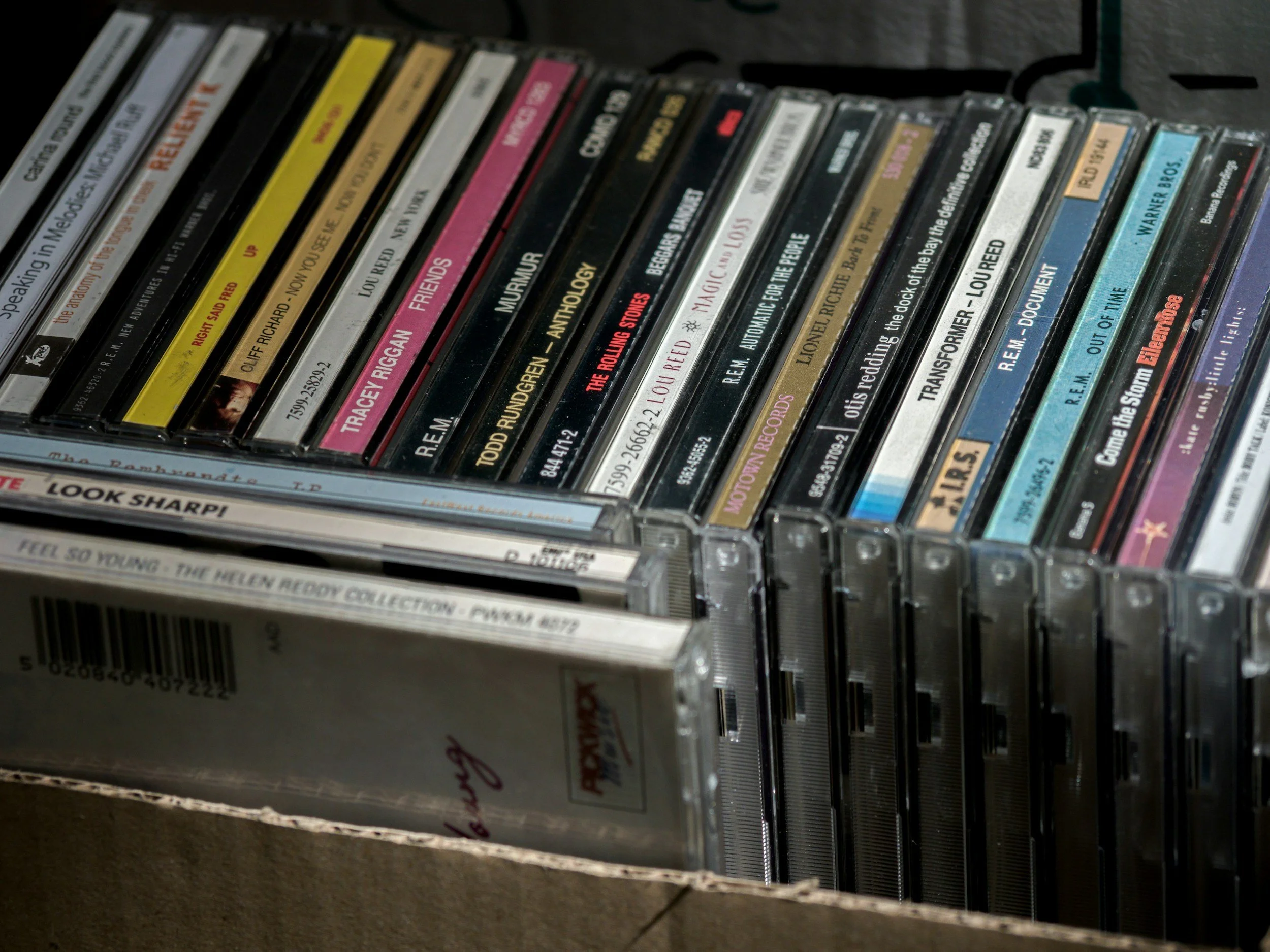 A collection of CD cases stored in a cardboard box, with some CD titles visible on the spines, such as 'Speaking in Melodies,' 'Reignite,' and 'Friends.'