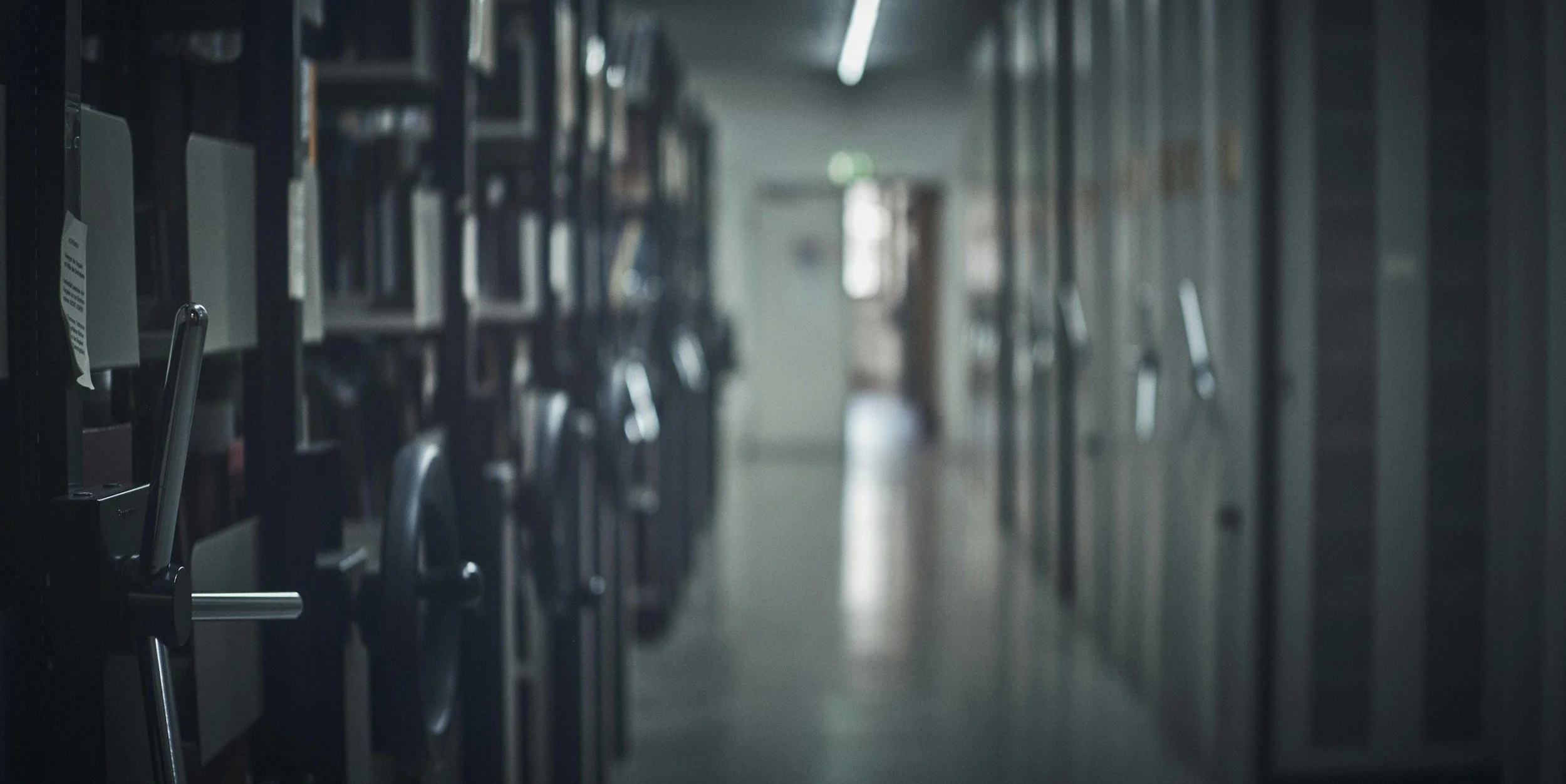 A long hallway with rows of archival shelving, with a blurred door at the end and soft natural lighting.