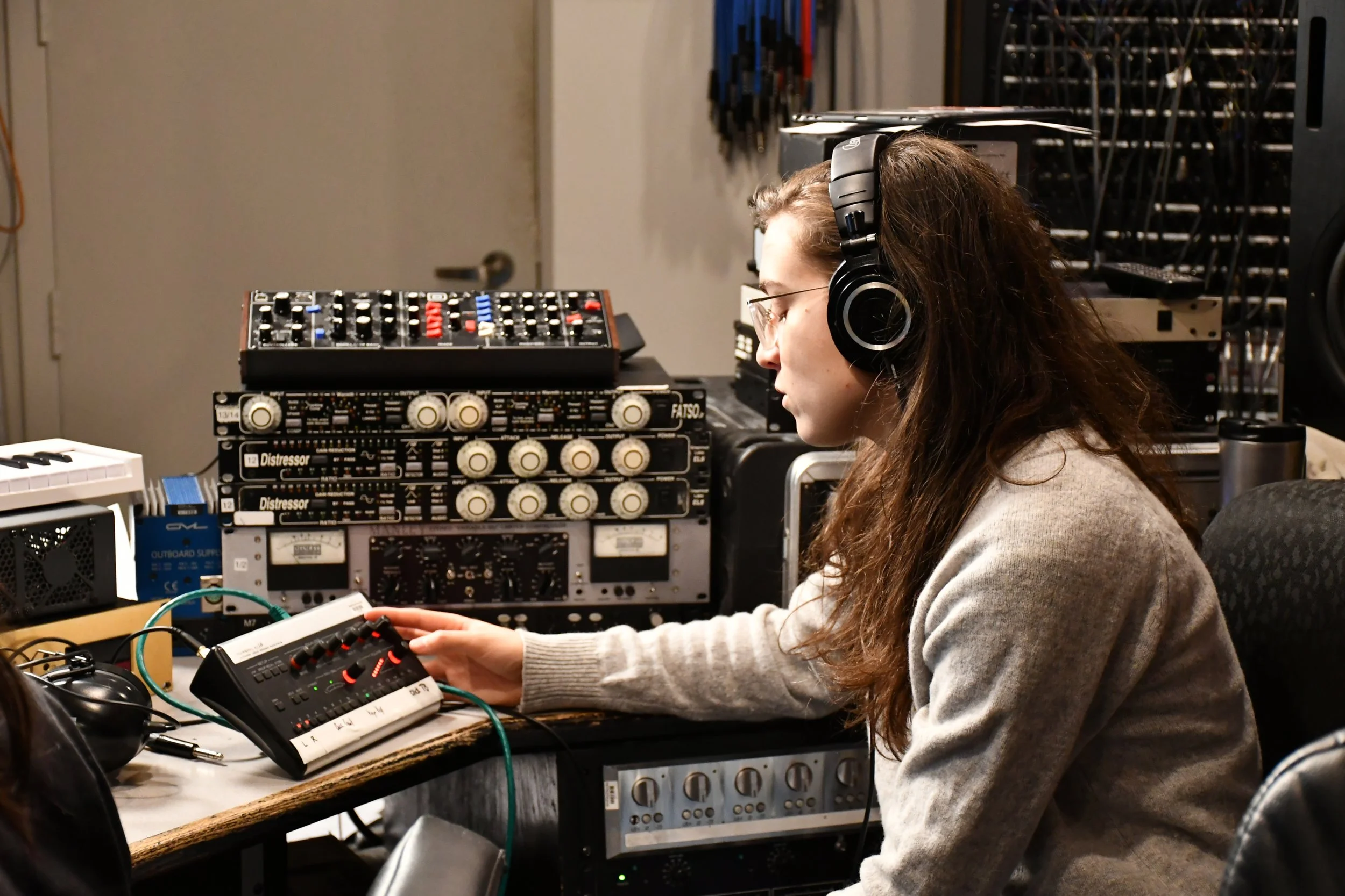 A woman with long brown hair, glasses, and a gray sweater wearing headphones, working with audio equipment in a studio.