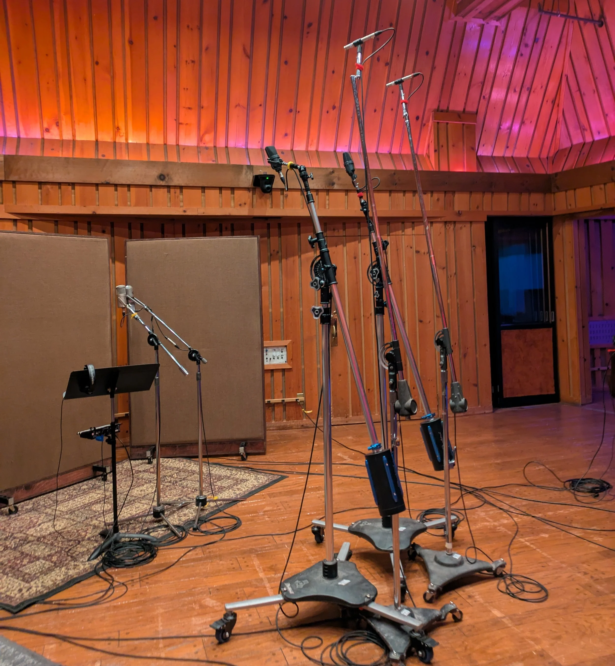 Recording studio with microphones, stands, and a music stand on a wooden floor, with sound panels and wooden wall in the background.