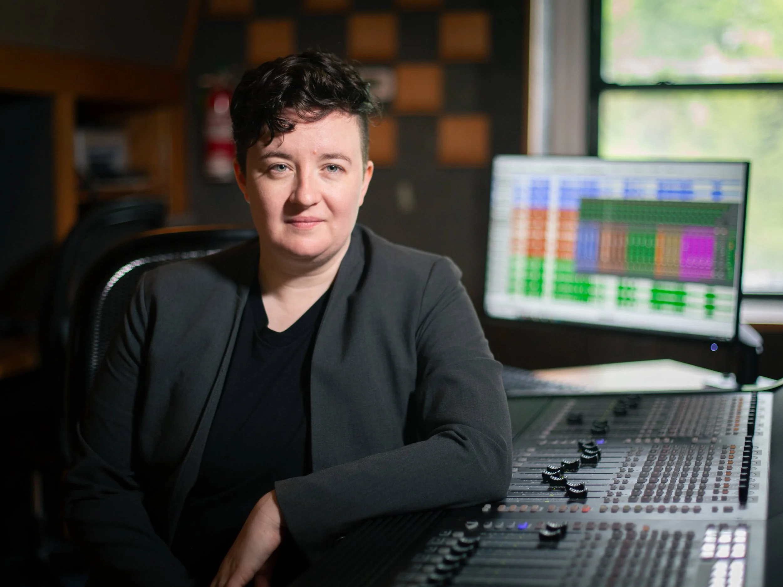 A woman sitting in a recording studio with a professional audio mixing console and a large computer monitor displaying a digital audio workstation.