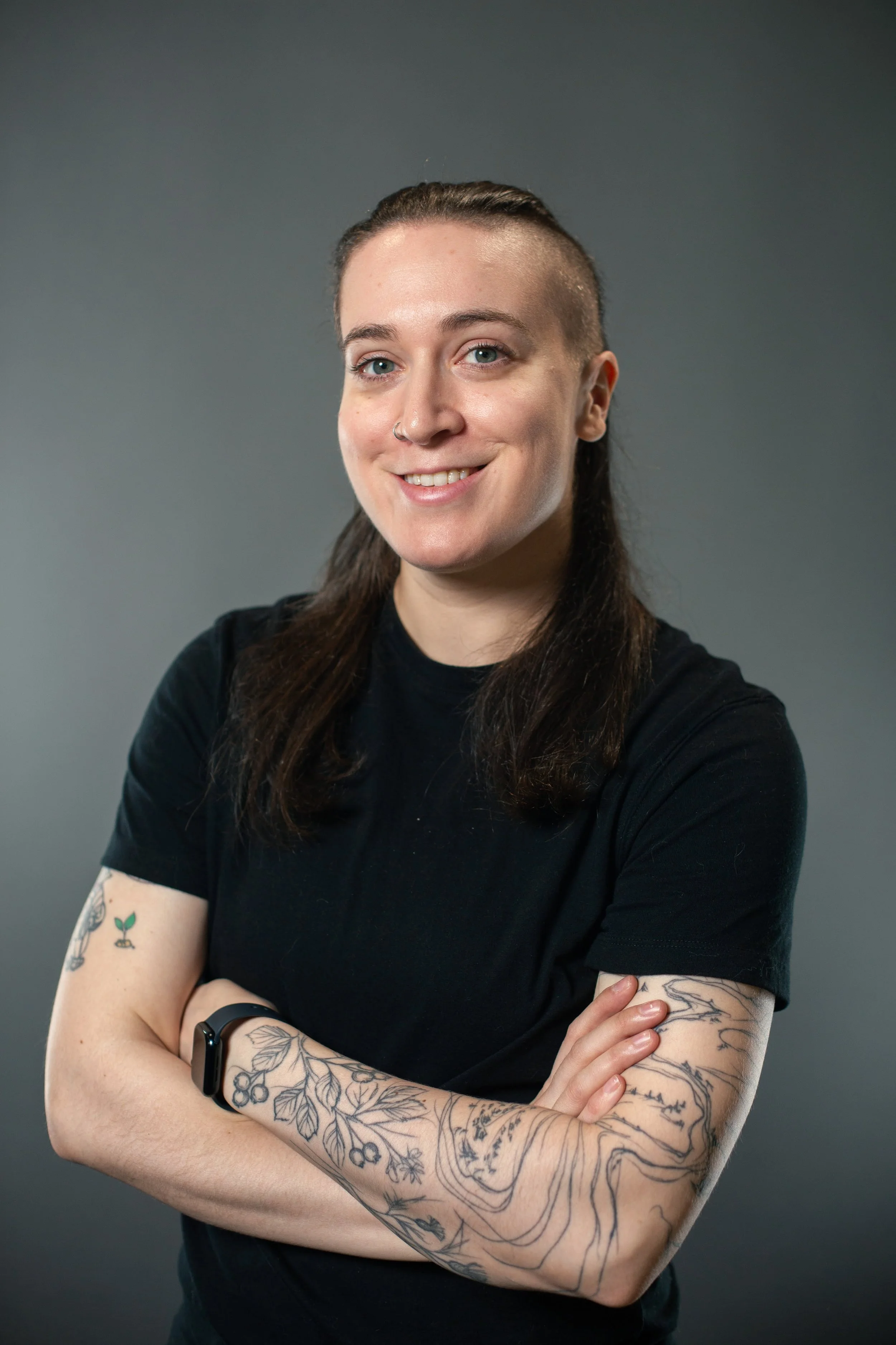 Portrait of a woman with tattoos on her arms, wearing a black t-shirt and a smartwatch, smiling and crossed arms, against a gray background.