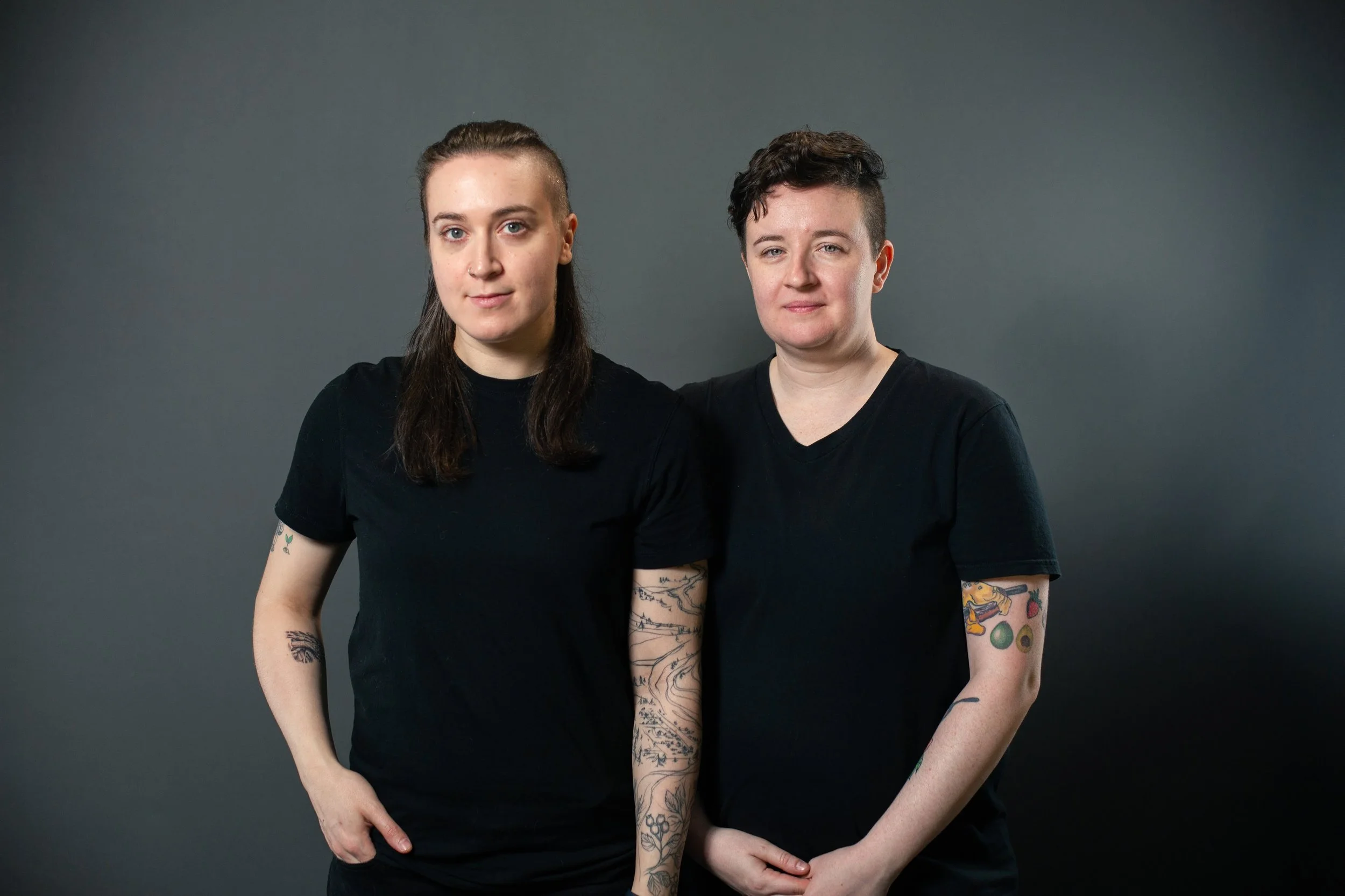 Two women with tattoos wearing black t-shirts standing side by side against a dark grey background.