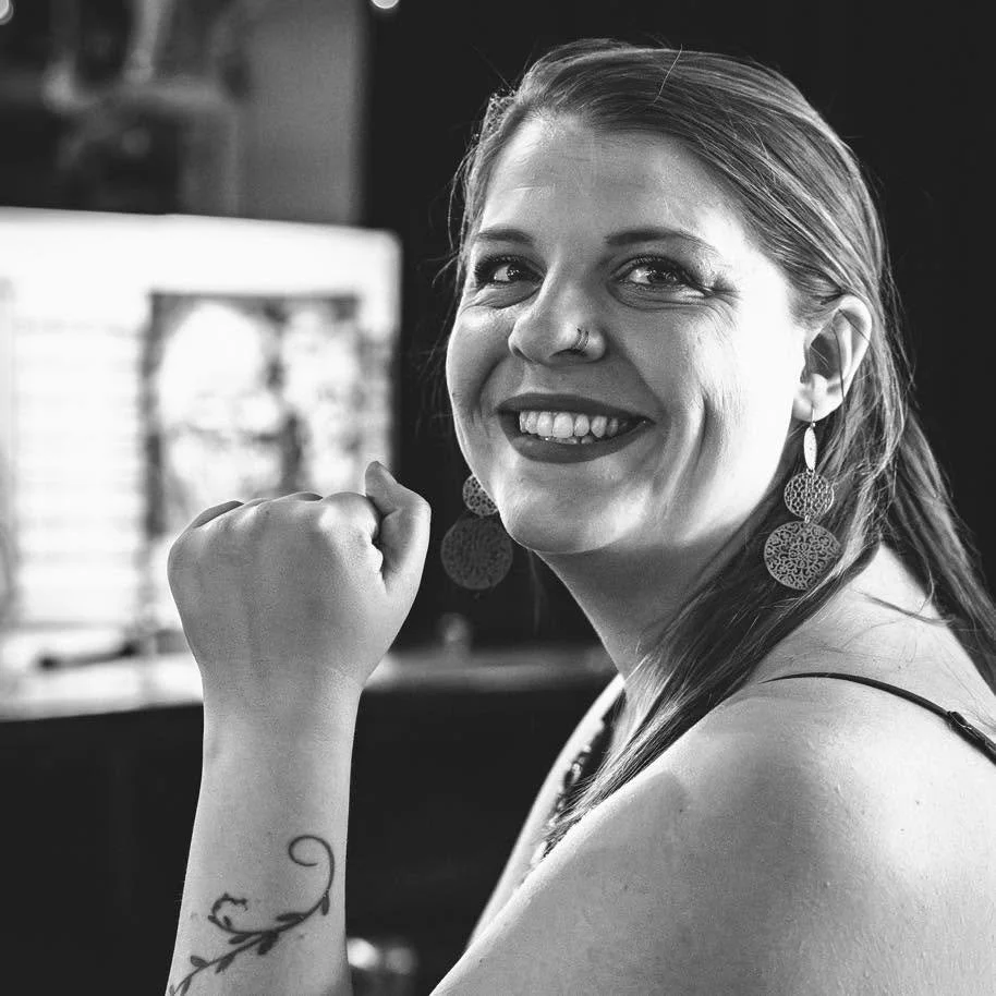 A smiling woman with earrings and a tattoo on her arm, holding her fist near her face, in black and white.
