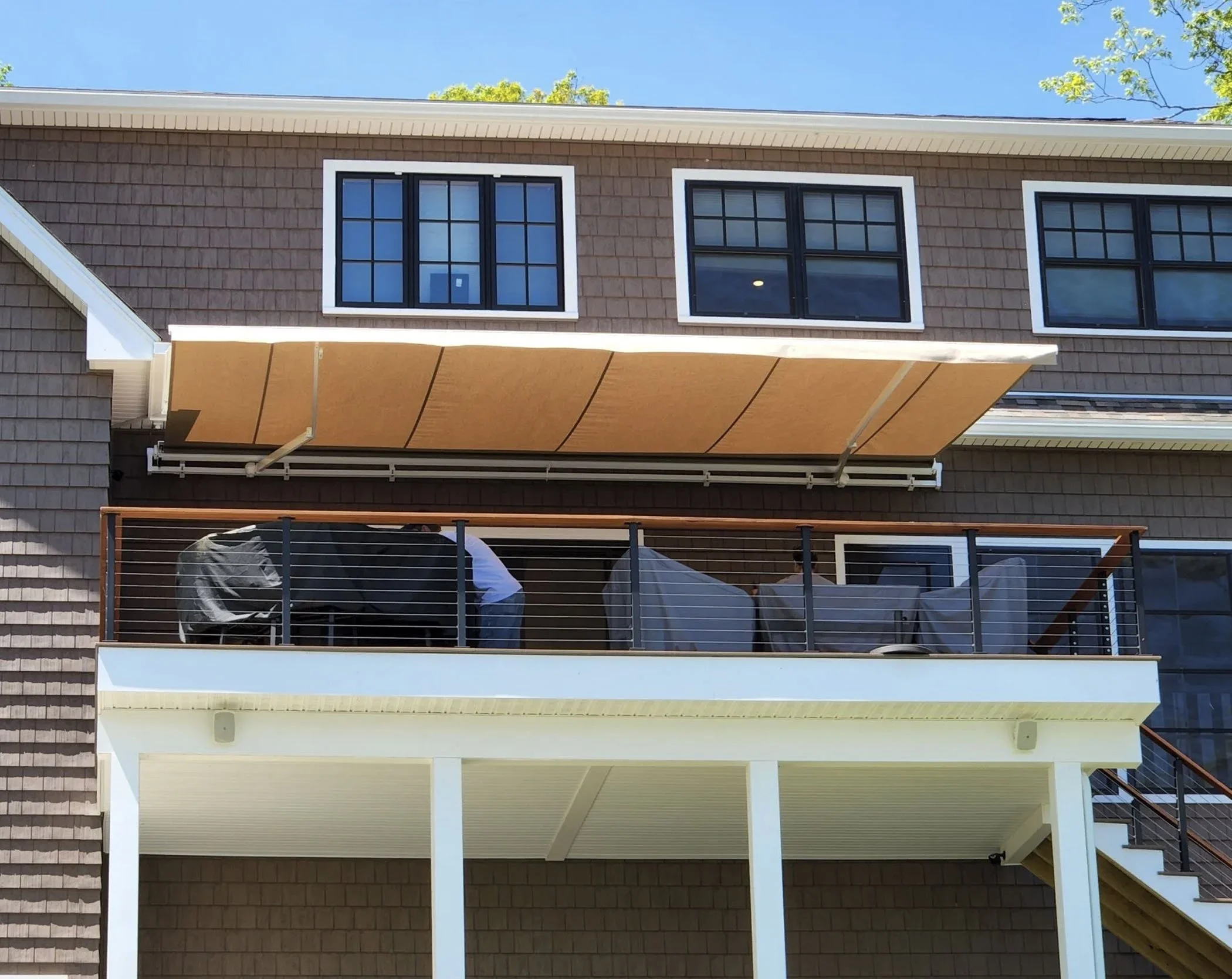 Multi-story house with brown shingle siding, three large windows on the upper floor, a beige retractable awning extending over a balcony, and outdoor furniture and a BBQ grill covered with a black cover on the balcony.