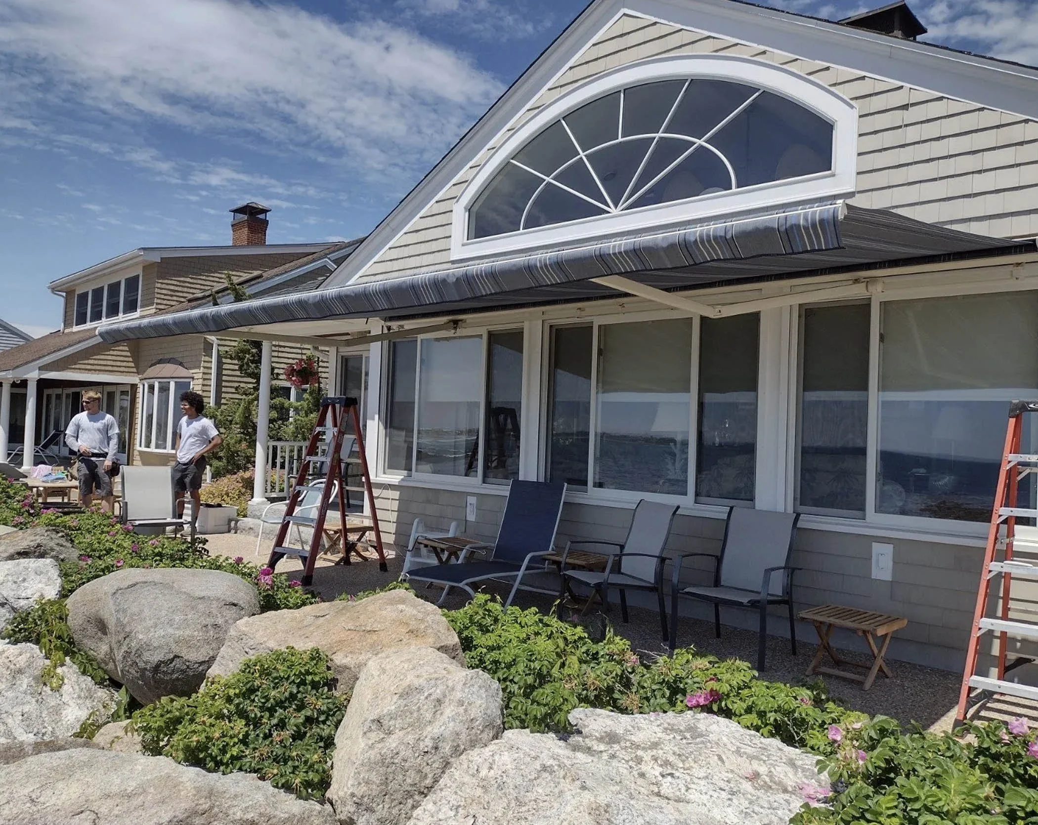 People working on a house exterior with ladders and outdoor chairs, rocks, and flowering bushes in front of the house under a blue sky.