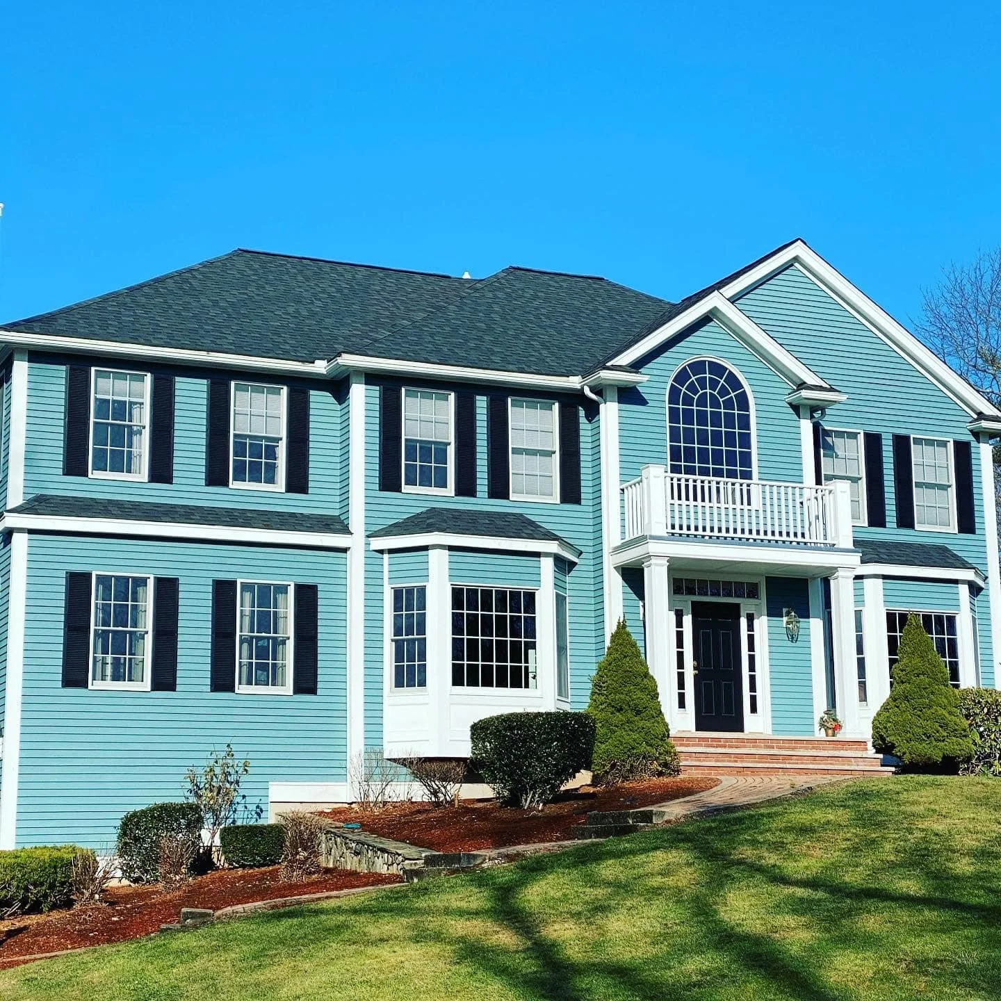A large, two-story house painted in light blue with white trim and a dark gray roof. The house has multiple windows with black shutters and a central entrance with a black door. There is a small balcony above the entrance with a large arched window a
