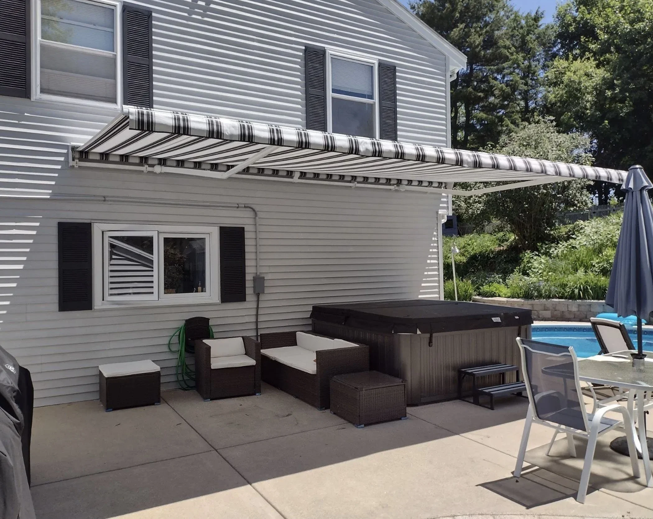 Backyard patio with a striped retractable awning, outdoor furniture including a cushioned seating area, a hot tub, a round table with chairs, an umbrella, and a swimming pool with blue water, surrounded by trees and greenery.
