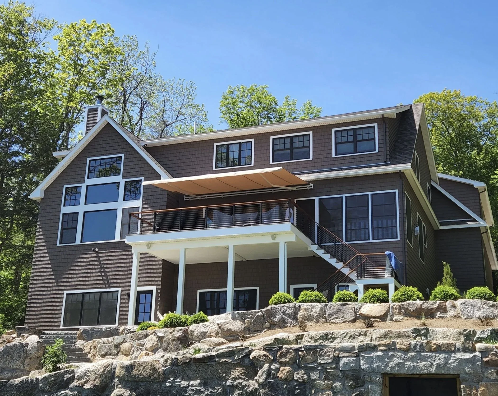 Large multi-story house with dark brown shingles, white trim, multiple windows, a balcony with a wooden railing, and an outdoor staircase, surrounded by trees and rocks under a clear blue sky.