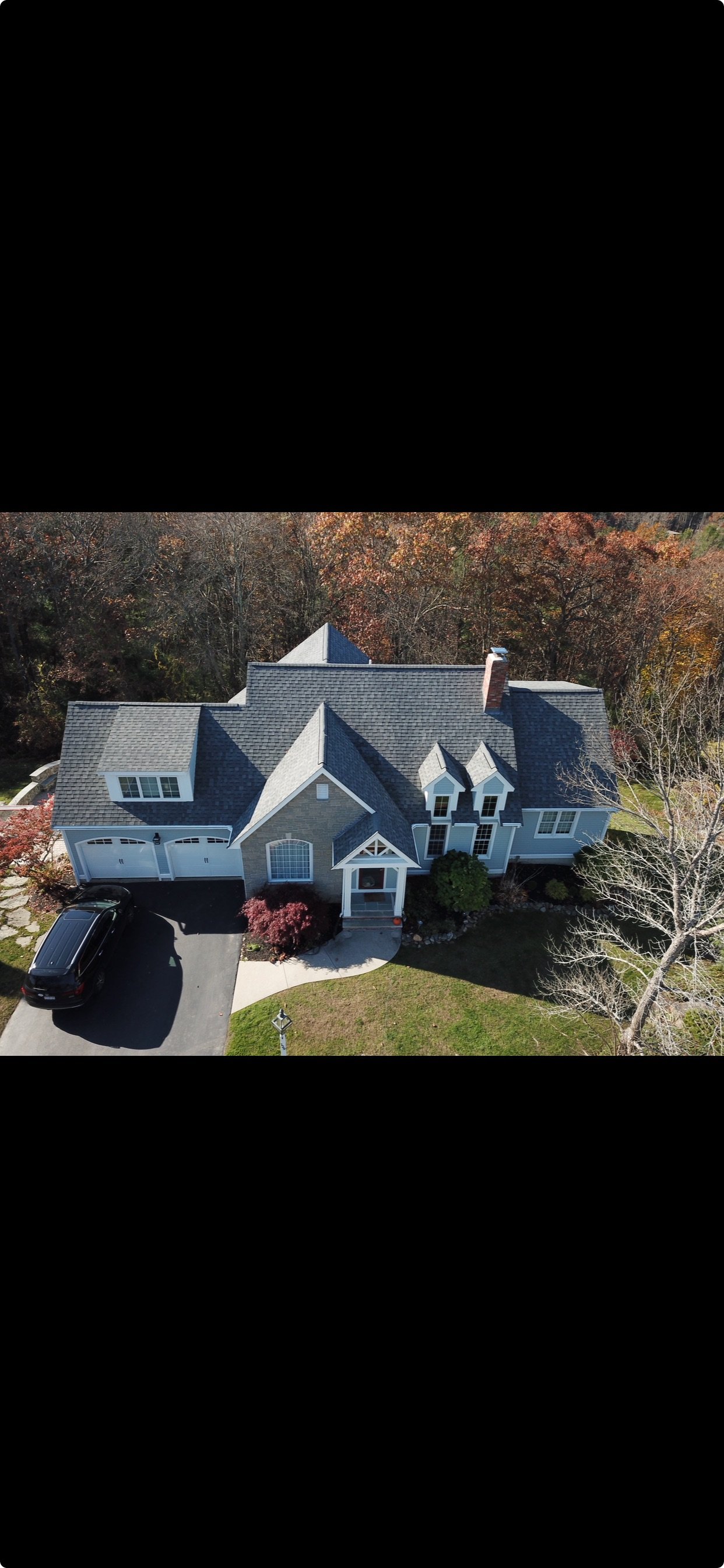An aerial view of a large, multi-story house with a gray roof, white windows, a garage with a black car parked in the driveway, and surrounded by trees with fall foliage.
