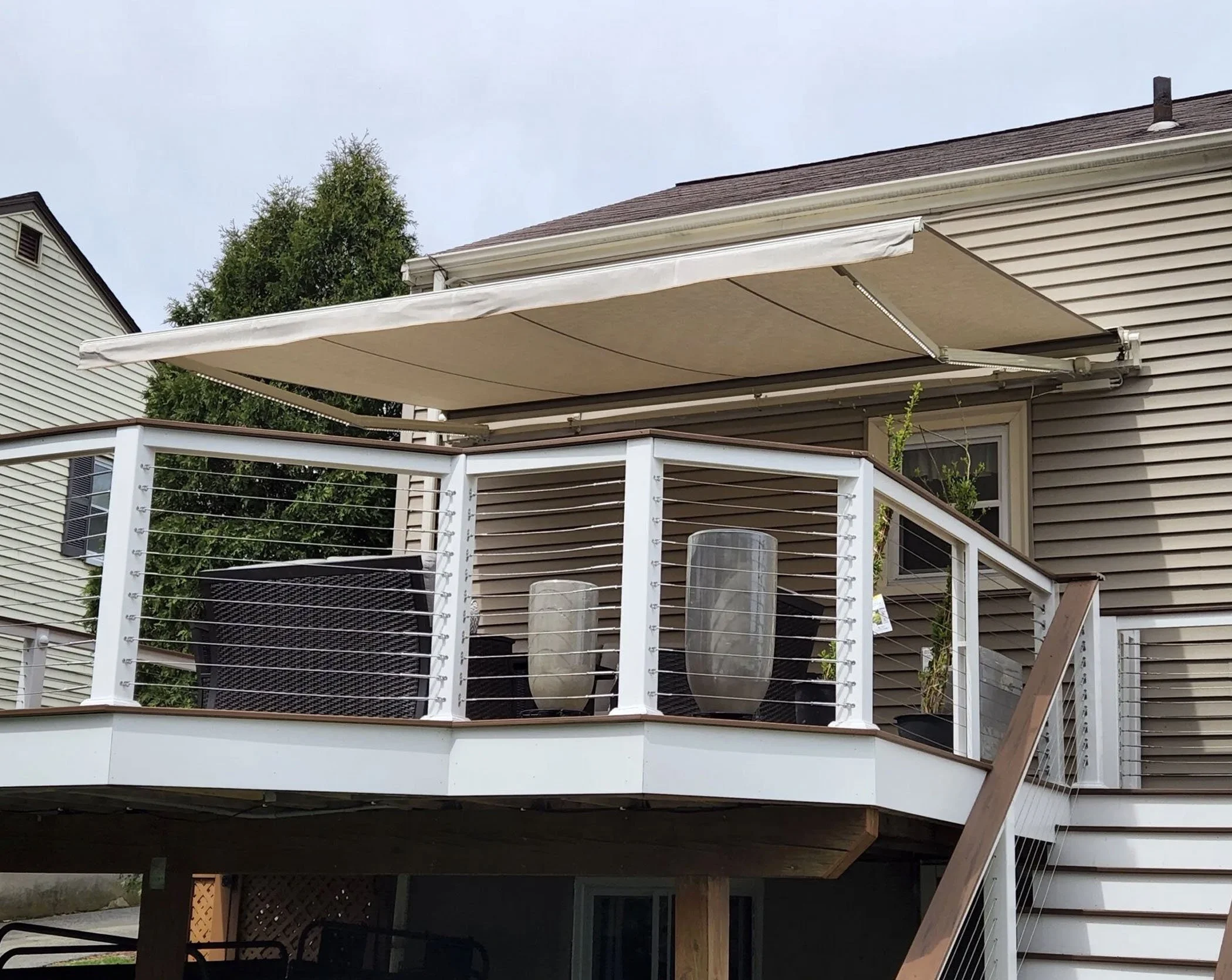 Residential house deck with white railing, outdoor furniture, potted plants, and a retractable beige awning, with neighboring house and trees in the background.