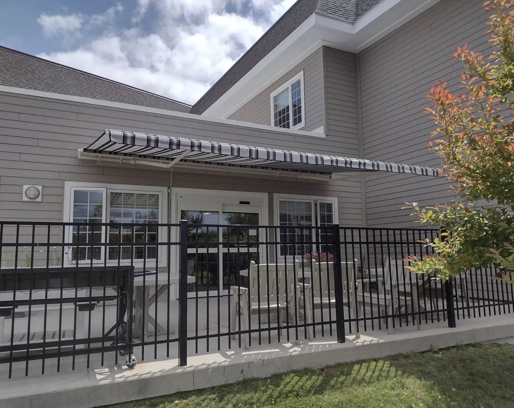Back patio of a house with a black metal fence, white outdoor furniture, and a striped awning above sliding glass doors, with a small tree on the right and partly cloudy sky overhead.