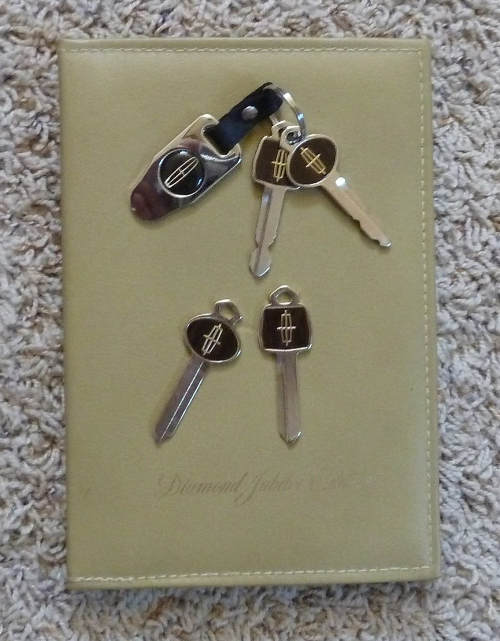 A set of five keys with keychains resting on a yellow book or journal on a beige carpet. The keys include a black car key, a house key, and three other keys. All keys have black keychain tags with a silver logo.